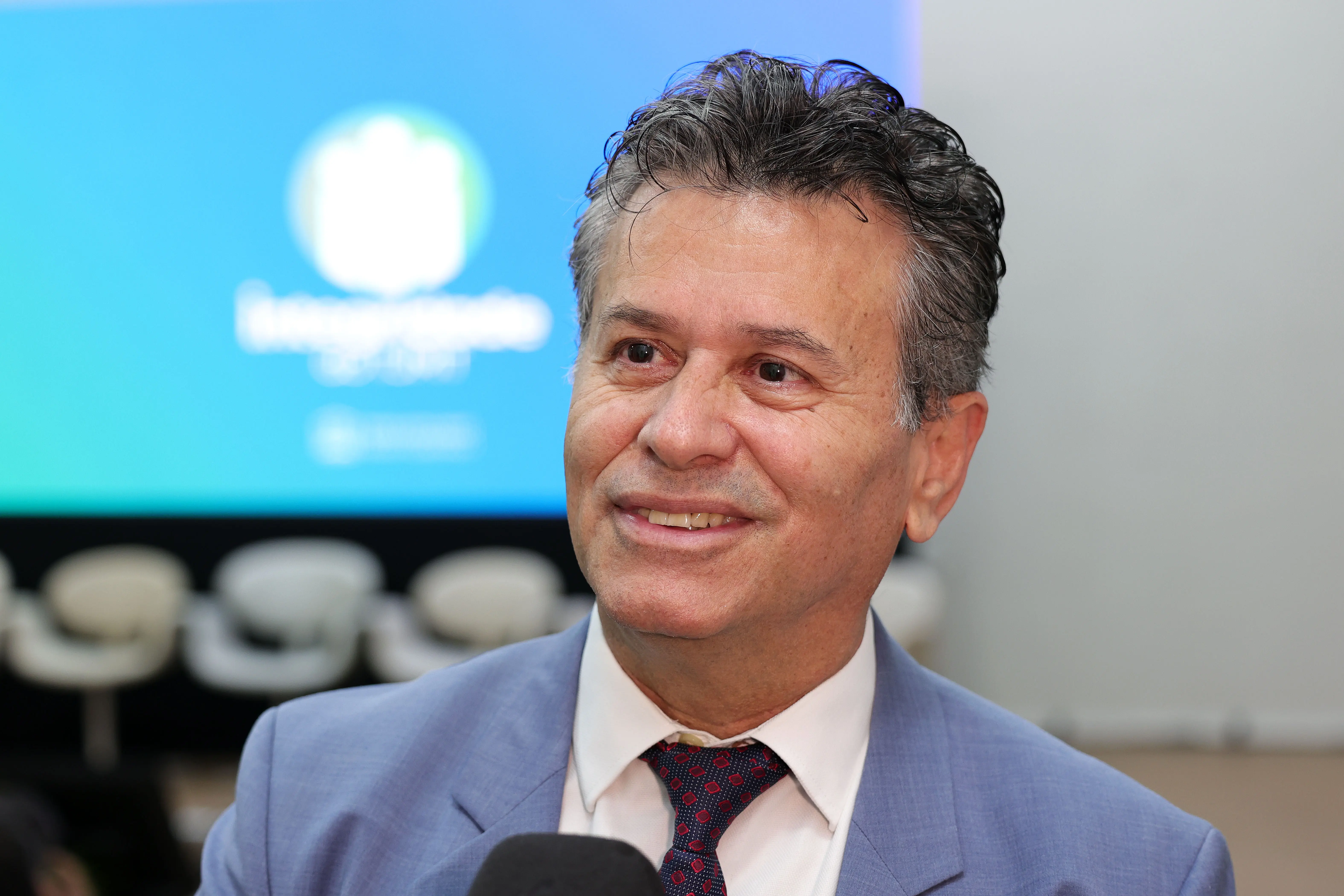 Foto do desembargador Jones Gattass Dias. Ele tem cabelos grisalhos curtos e bem penteados, sorri e olha para a direita. Ele veste um terno azul claro, camisa branca e gravata estampada. O fundo mostra cadeiras e uma tela.