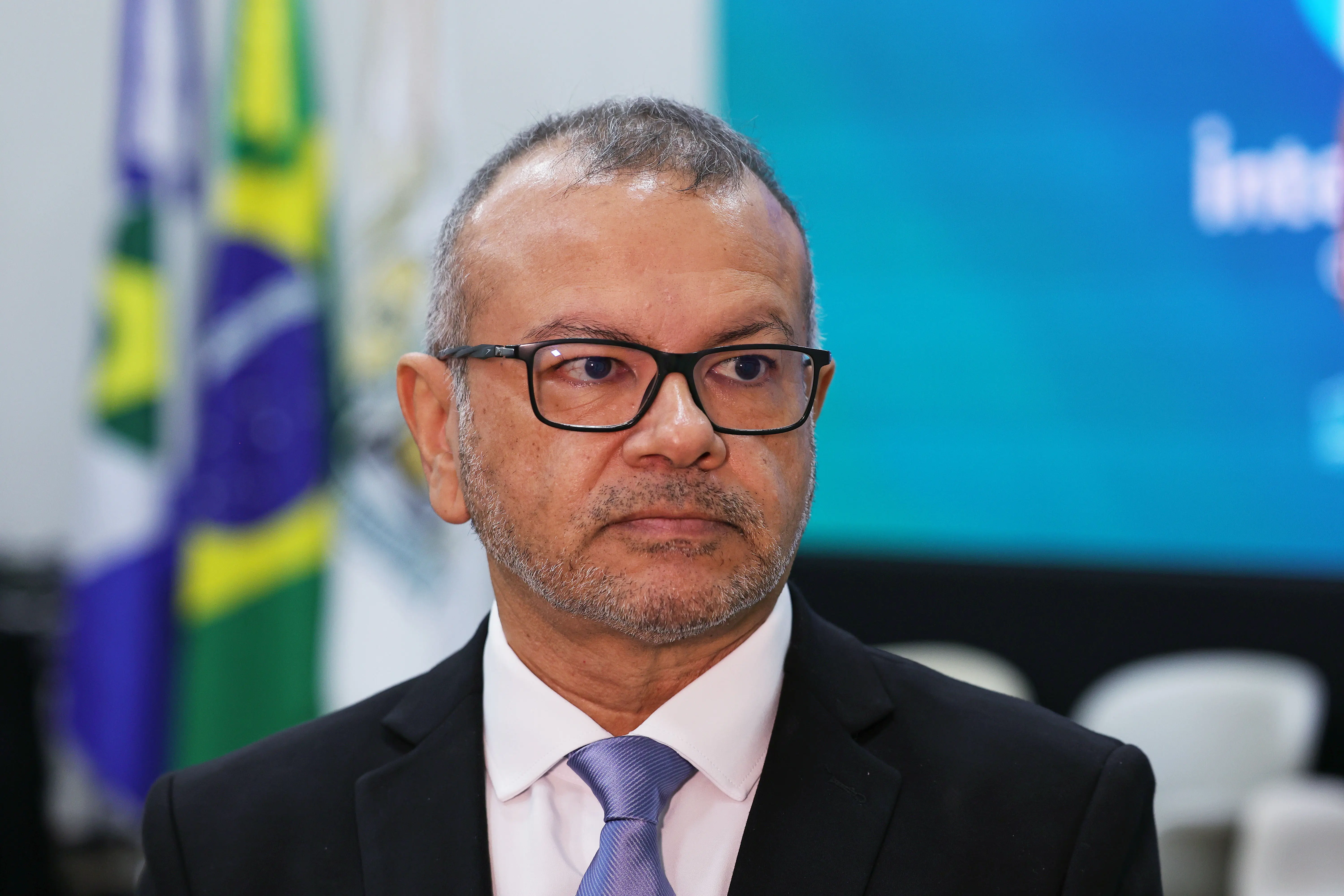 Retrato do juiz Alex Nunes de Figueiredo. Ele tem  pele morena, usa óculos de grau com armação preta e barba rala. Veste um terno preto, camisa branca e gravata roxa, com uma expressão neutra e olhar direcionado para a direita. Bandeiras estão desfocadas ao fundo.