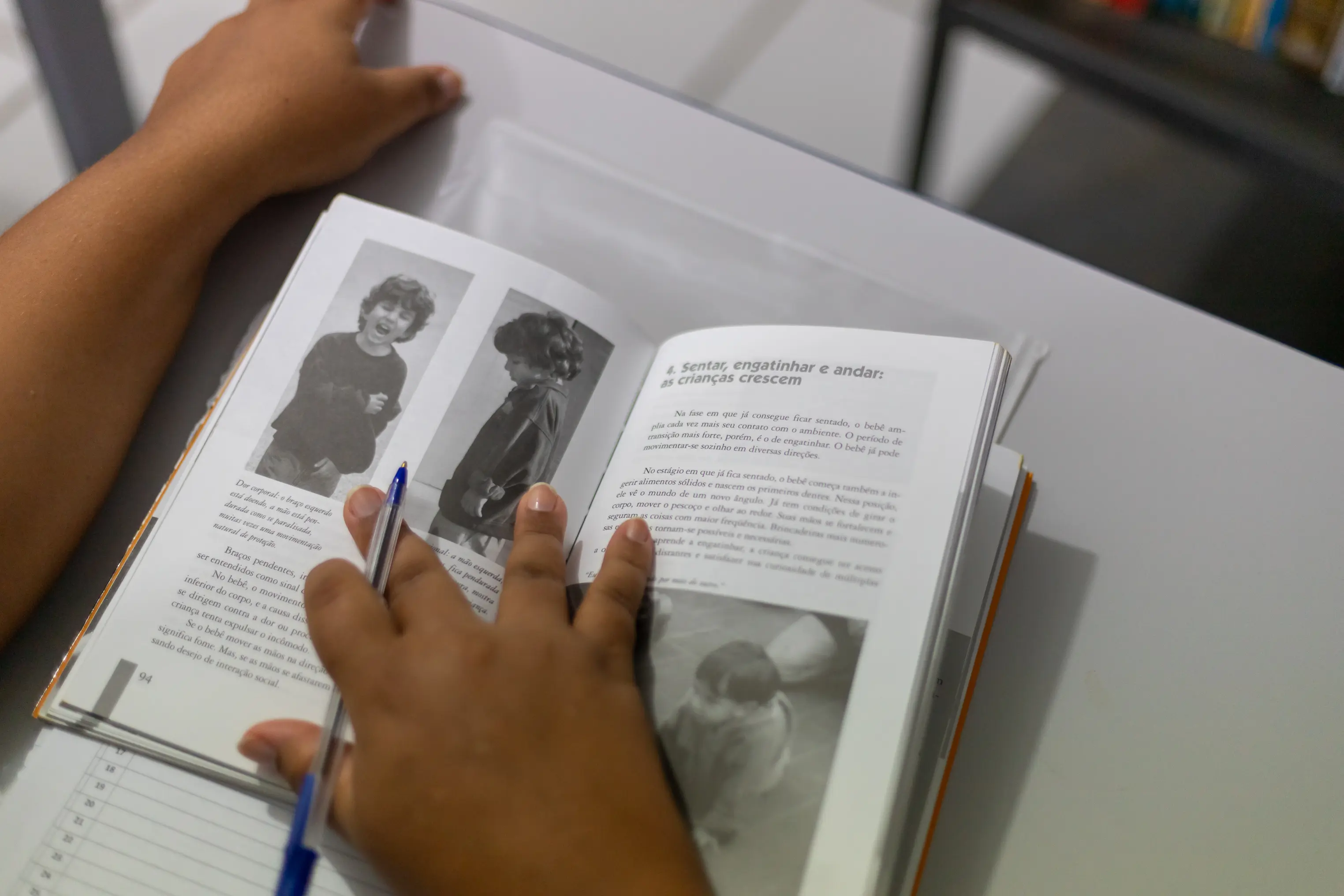 Visão em ângulo de uma pessoa folheando um livro aberto sobre uma mesa branca. Uma das mãos segura uma caneta azul, apontando para o texto que traz fotos em preto e branco de crianças.