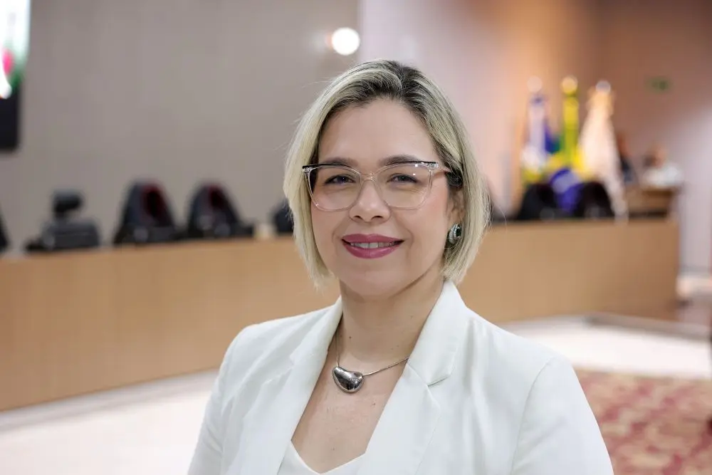 Foto horizontal que mostra a assessora do desembargador Sérgio Valerio, Evaneide Amaral, do busto pra cima, sorrindo para a foto, no plenário do TJMT. Ela é uma mulher branca, de cabelo chanel loiro, usando óculos de grau e blazer branco.