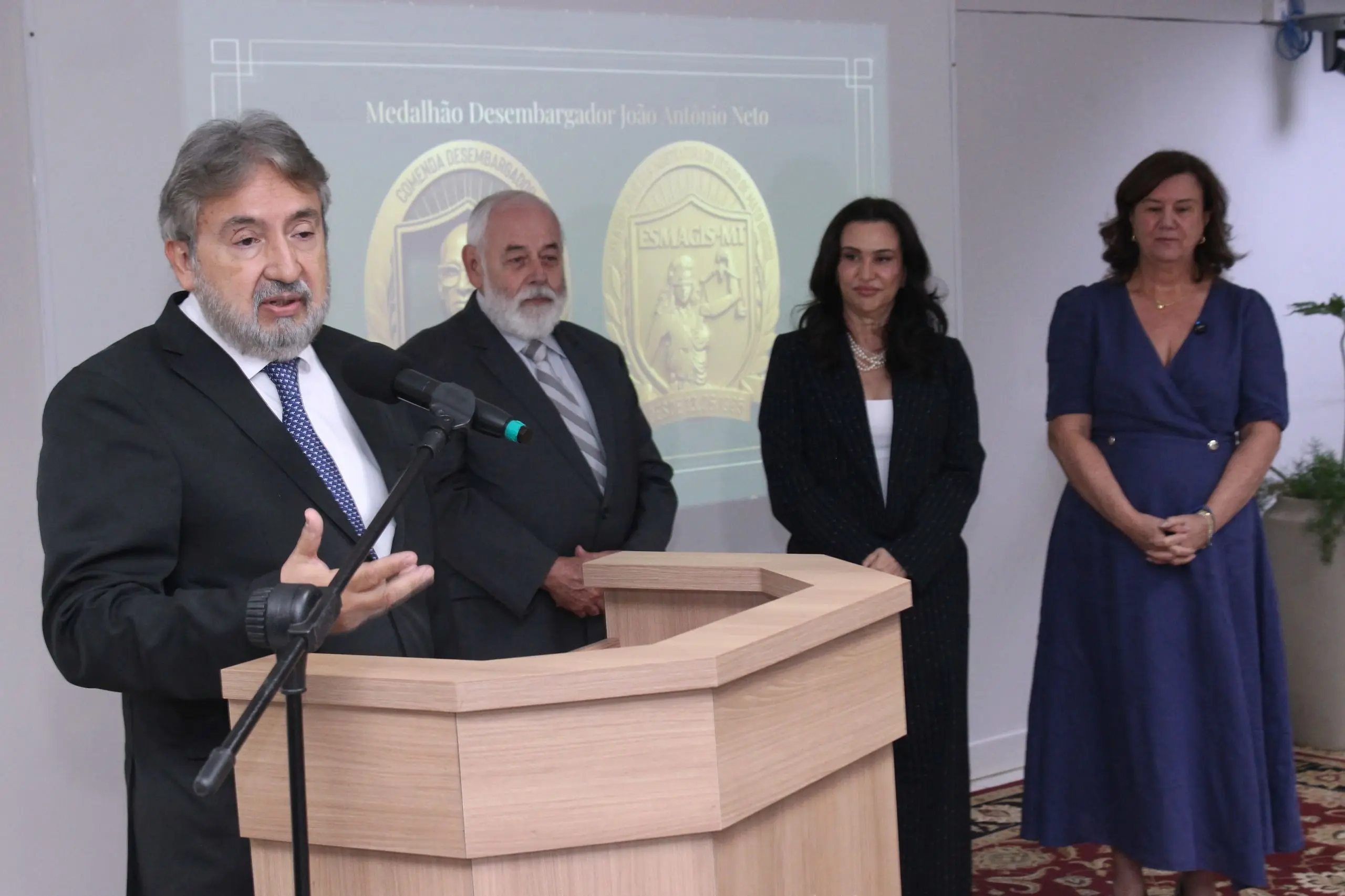 Homem de terno preto discursa em um púlpito à esquerda. Atrás dele, um homem e duas mulheres observam em pé. Ao fundo, uma projeção mostra a imagem de duas medalhas douradas da Esmagis-MT.