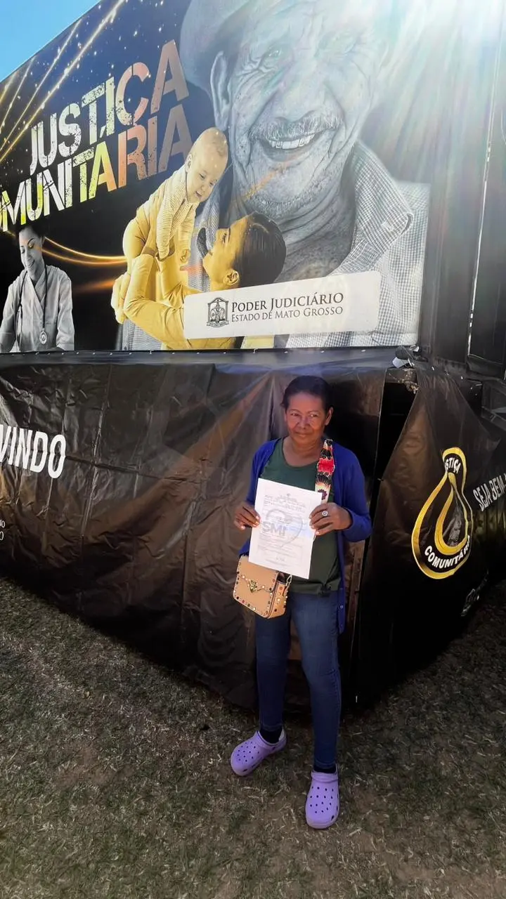 Foto vertical que mostra a aposentada Maria Rita do Carmo Freitas em pé, mostrando um papel, em frente à carreta oftalmológica da Justiça Comunitária. 