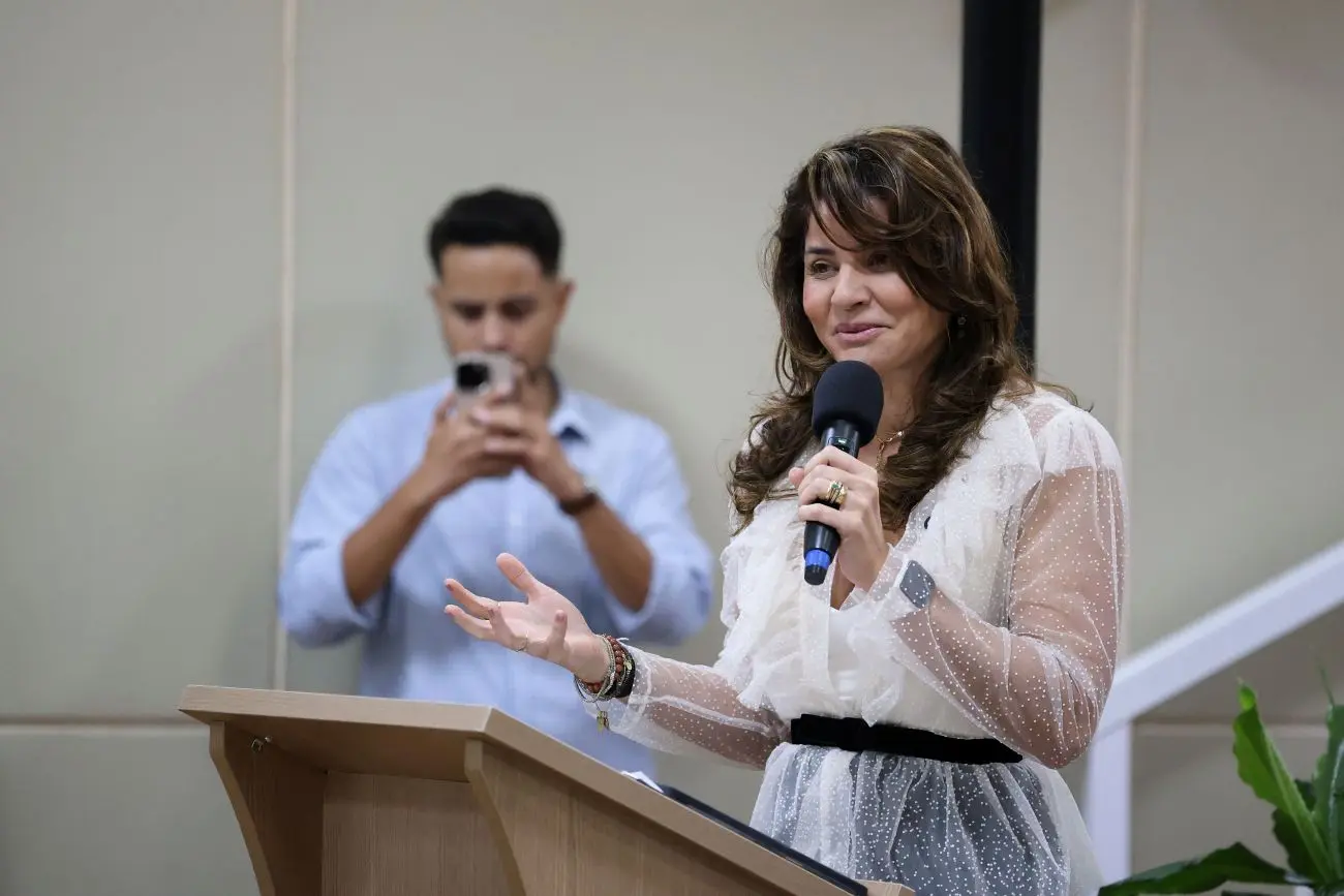 Foto horizontal que mostra a defensora-pública geral, Luziane Castro, falando ao microfone, no púlpito do auditório. Ela é uma mulher de pele clara, longos cabelos castanhos e ondulados, usando saia preta, blusa branca e capa transparente de tule branco, com franjas na gola e cinto preto.