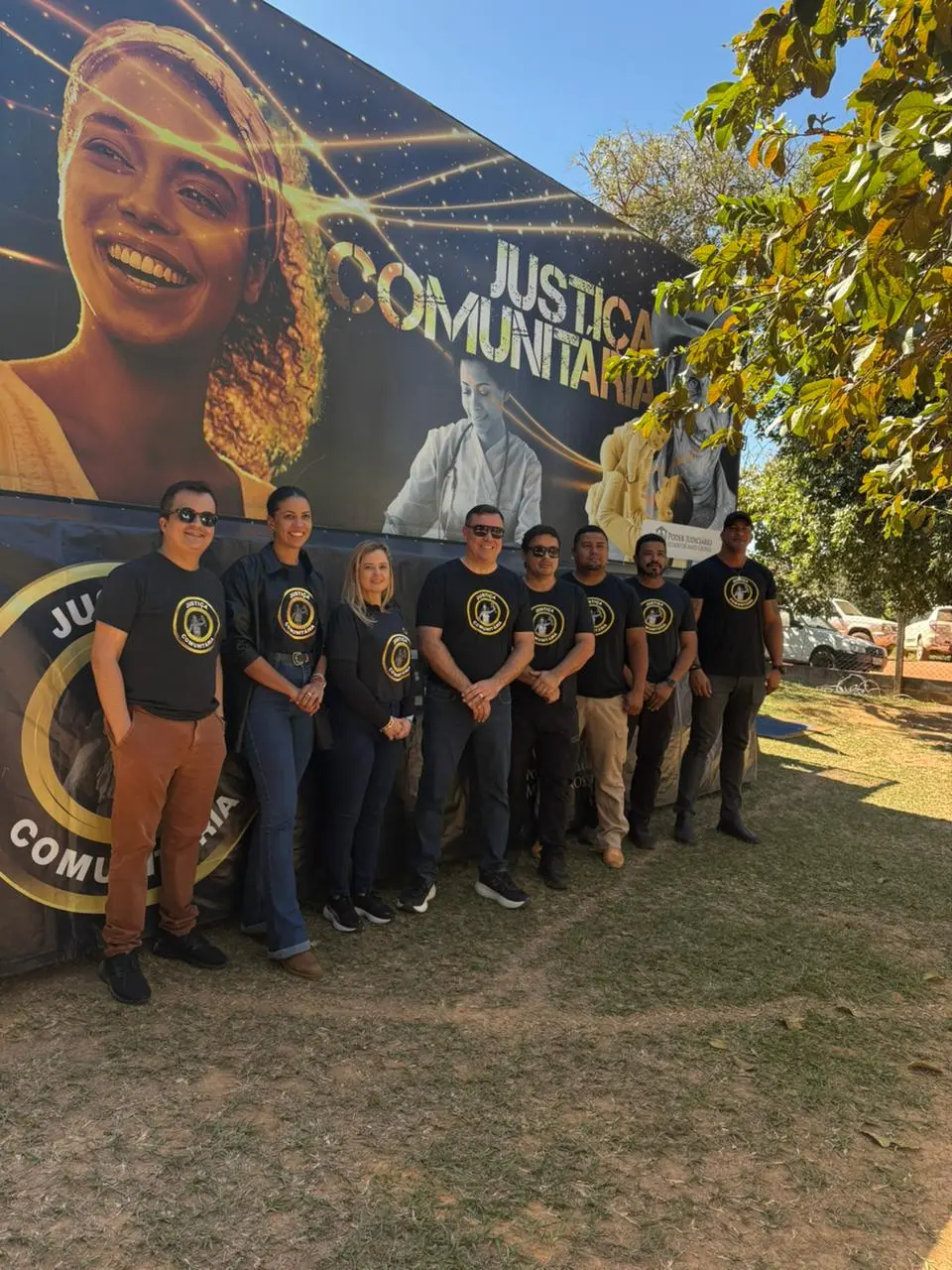 Foto vertical que mostra oito pessoas, sendo seis homens e duas mulheres, usando camiseta preta com logomarca da Justiça Comunitária, posando em pé para a foto, em frente à carreta da Justiça Comunitária, em um campo aberto com grama e árvores. 