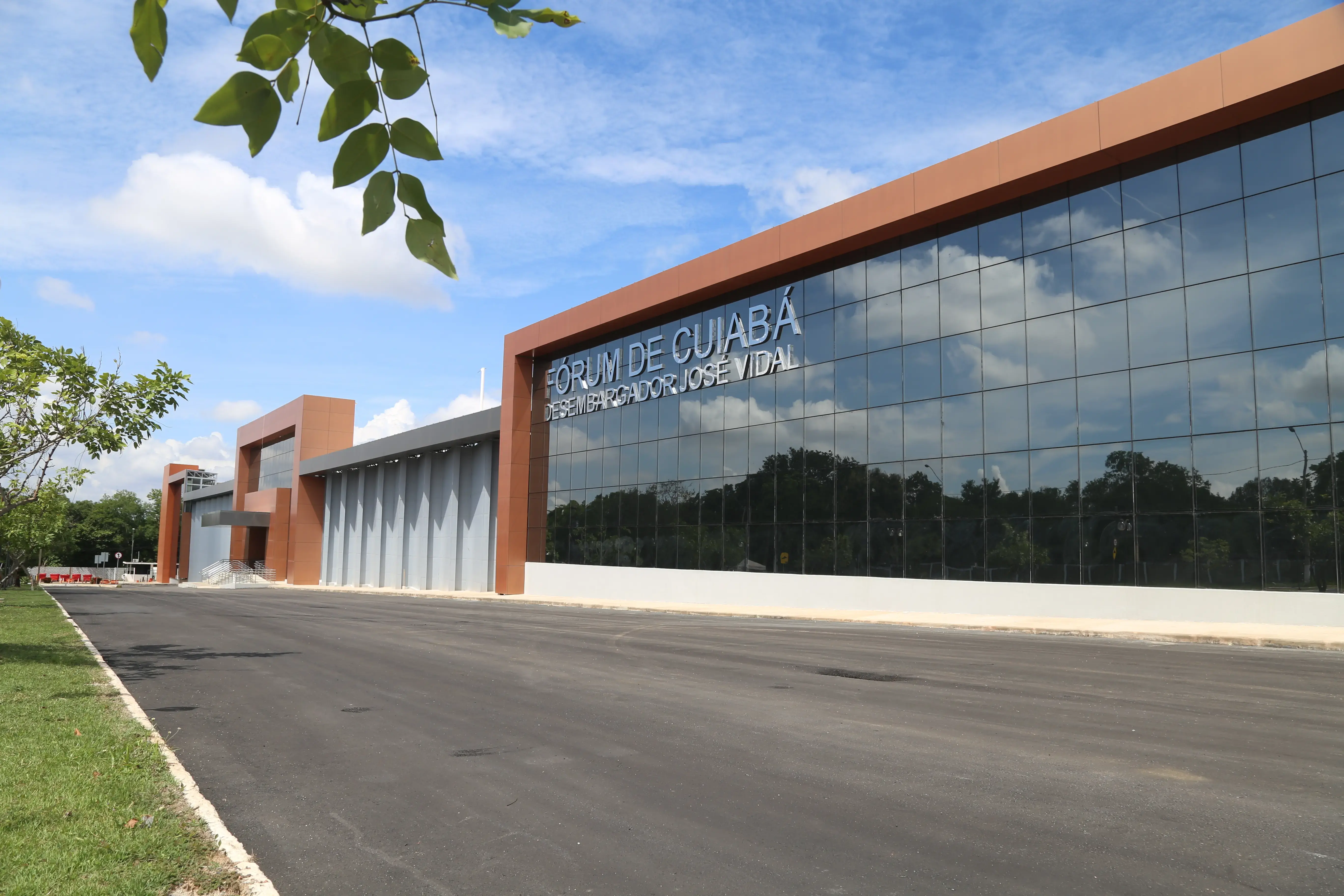 Fachada moderna do Fórum de Cuiabá sob céu azul com nuvens. O prédio longo tem uma grande parede de vidro espelhado que reflete as árvores, moldura em tom bronze e letreiro prateado na frente.