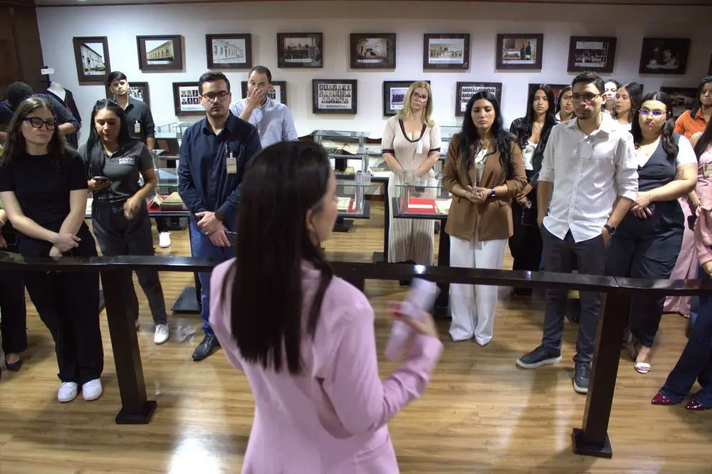 Foto horizontal em plano aberto que mostra um grupo de estudantes de Direito observando uma mulher de blazer cor-de-rosa, que está à frente deles, falando. Eles estão no Espaço Memória do Tribunal de Justiça de Mato Grosso, local cheio de quadros e mesas de vidros com documentos antigos expostos.
