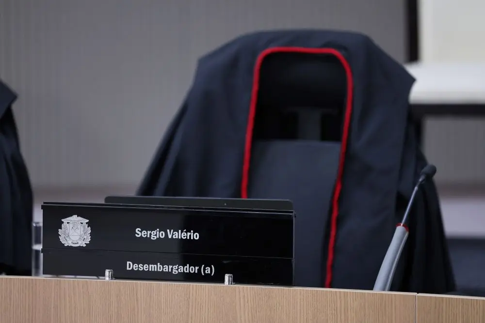 Foto horizontal que mostra a cadeira do desembargador Sérgio Valerio no plenário do TJMT, com sua toga no encosto. à Frente, sobre a mesa, a placa com o nome do desembargador.
