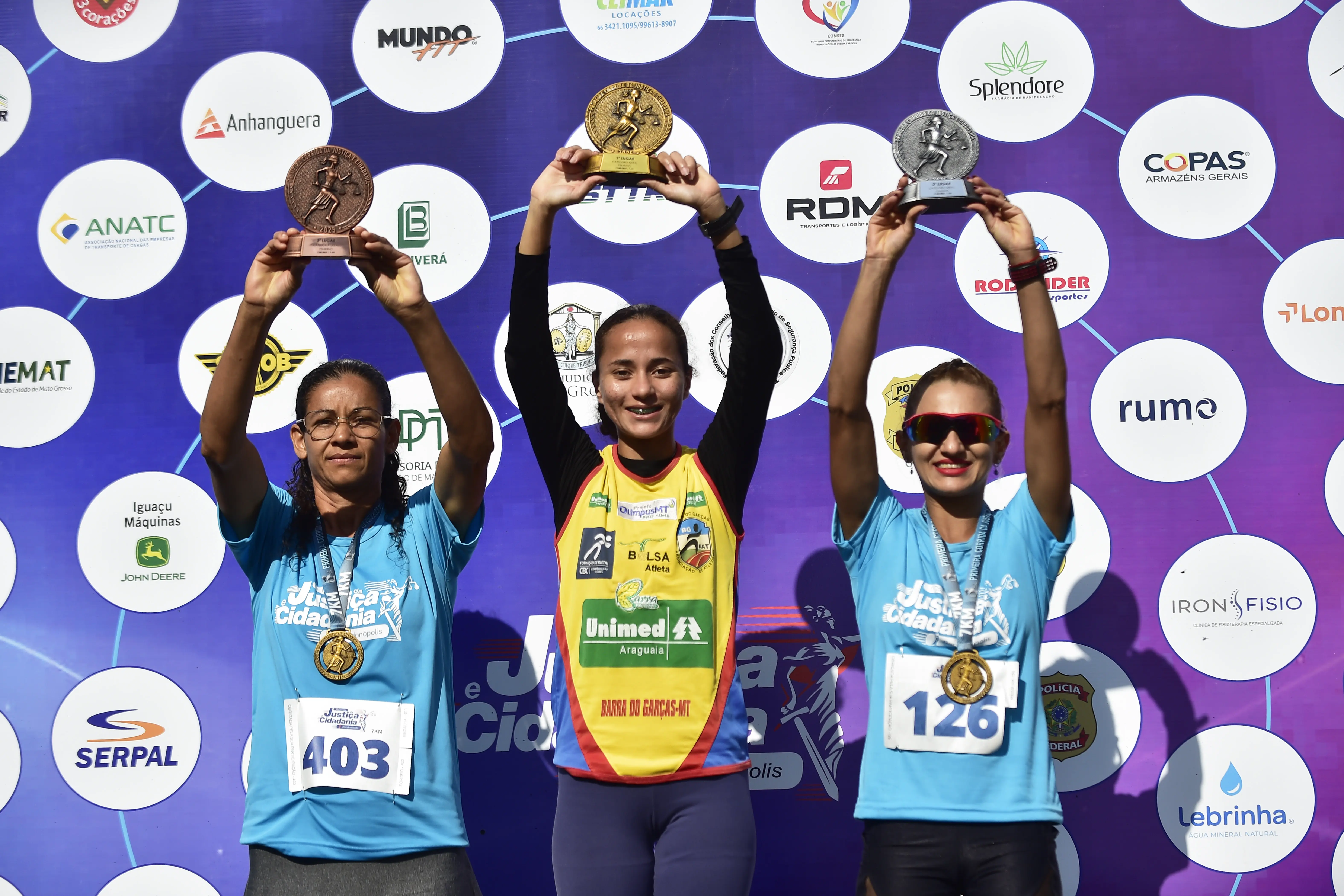 Três corredoras no pódio erguem seus troféus. A do centro veste camiseta amarela e as das pontas, azul-celeste com medalhas no peito. Ao fundo, um painel roxo com logomarcas de patrocinadores.