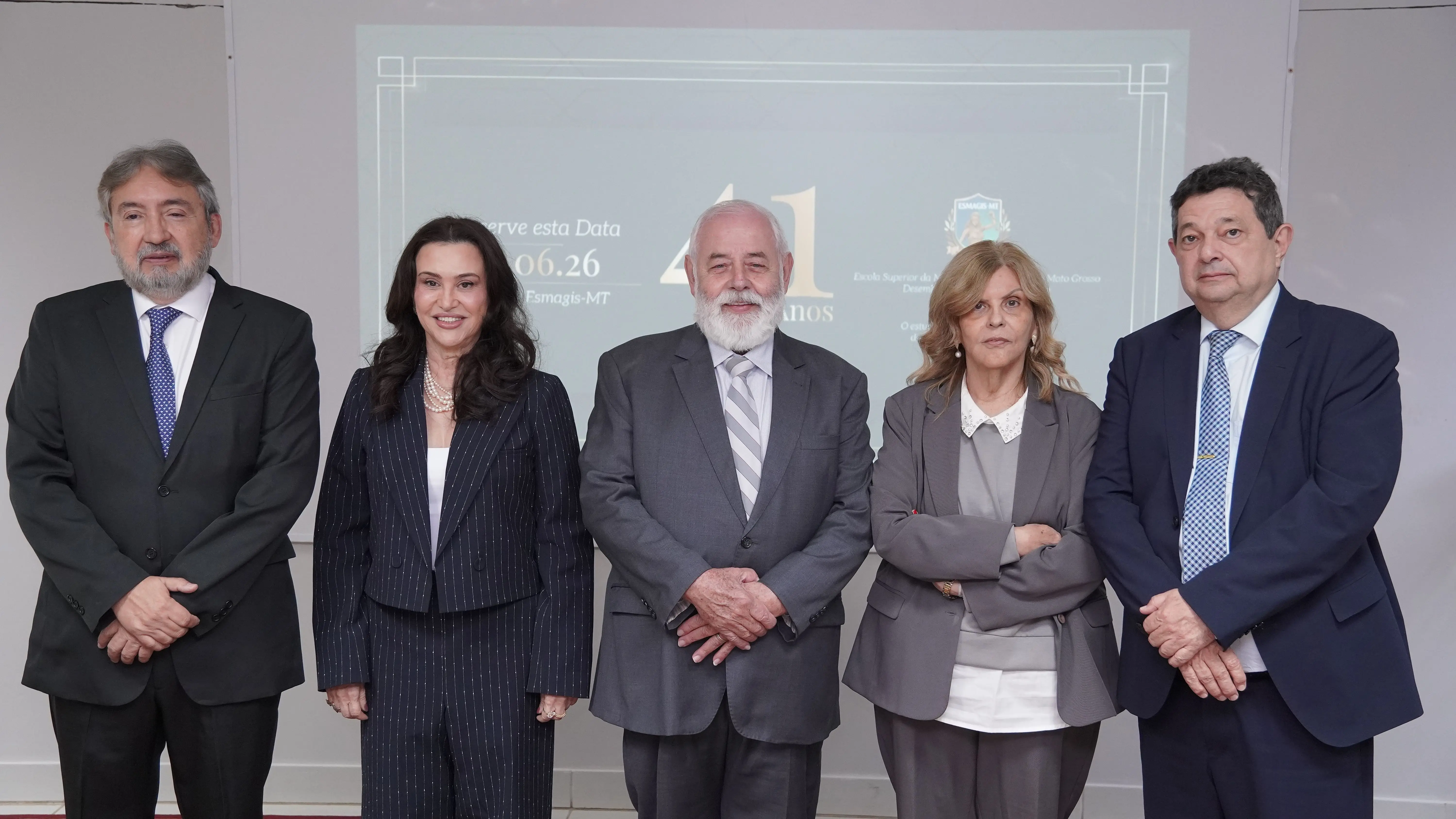 Cinco autoridades posam alinhadas lado a lado em um ambiente interno. Ao fundo, uma tela de projeção exibe o slide esverdeado de comemoração dos 