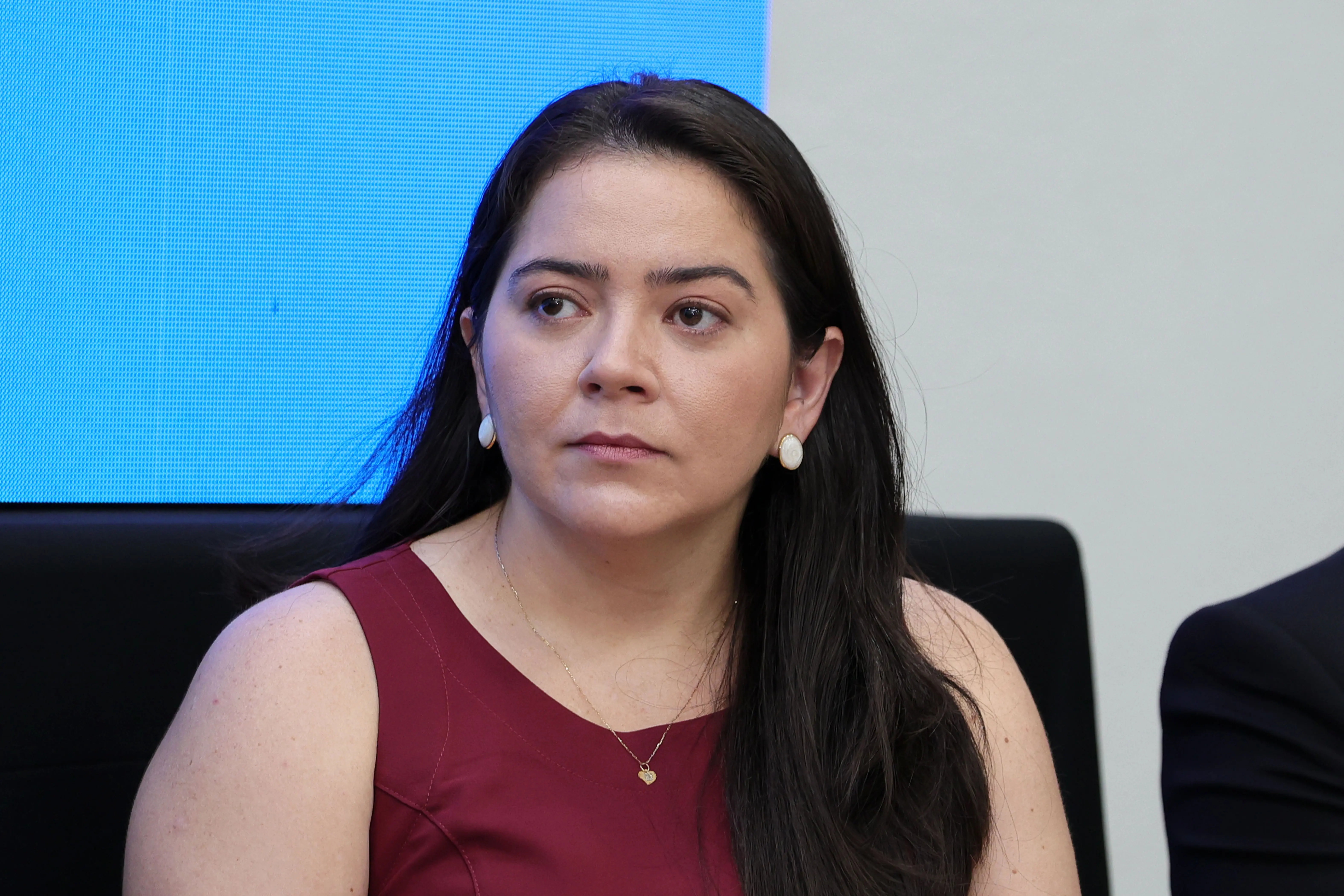 Retrato da controladora-geral da Defensoria Pública, Bárbara Nathalia Nogueira. Ela é uma mulher de pele branca e cabelos escuros, veste um vestido sem mangas na cor vinho e brincos pequenos. Ela olha levemente para a direita da imagem, com uma expressão séria. O fundo é desfocado.