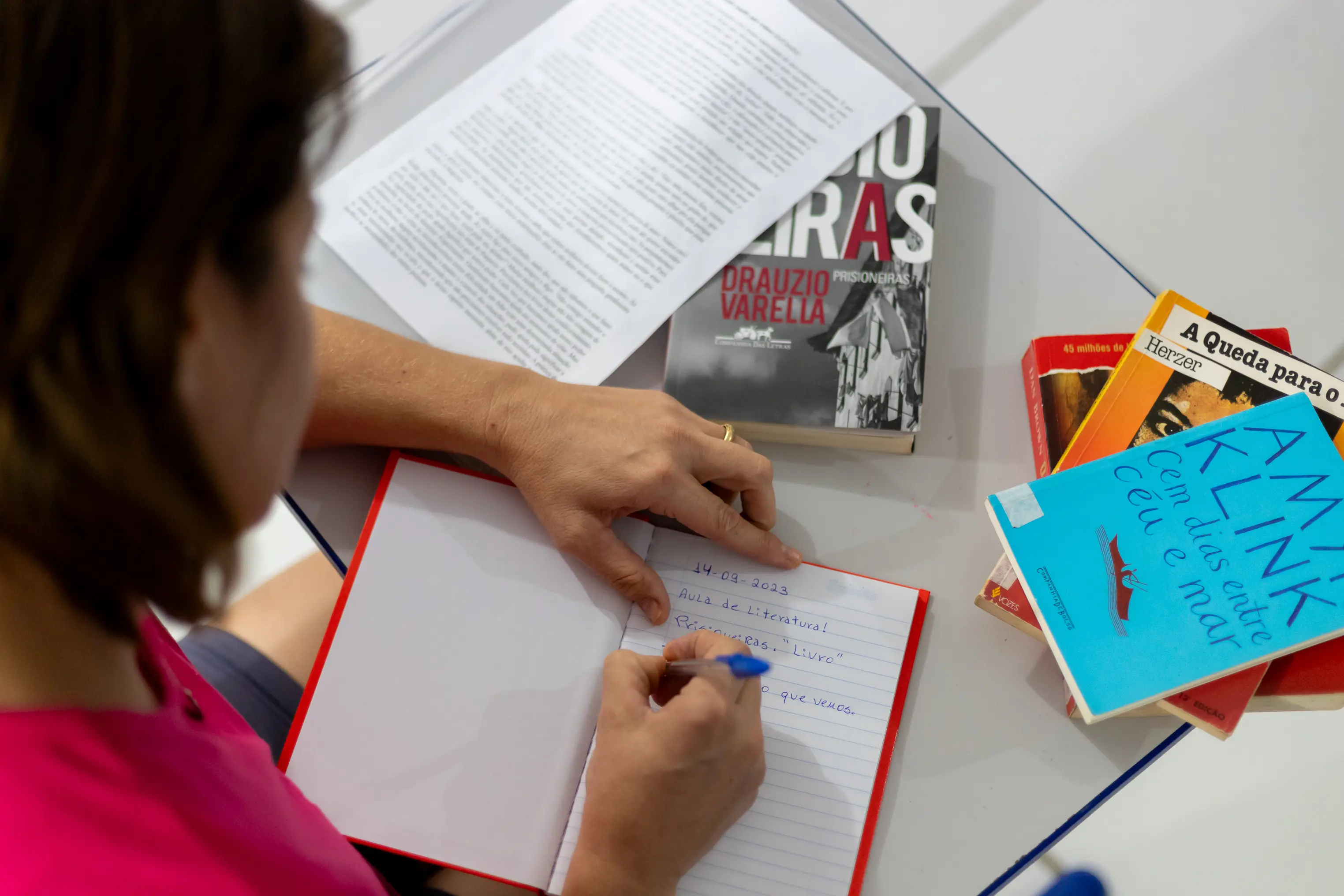 Vista de cima, uma mulher de blusa rosa escreve em um caderno de capa vermelha. Na mesa de vidro, há folhas impressas e os livros 