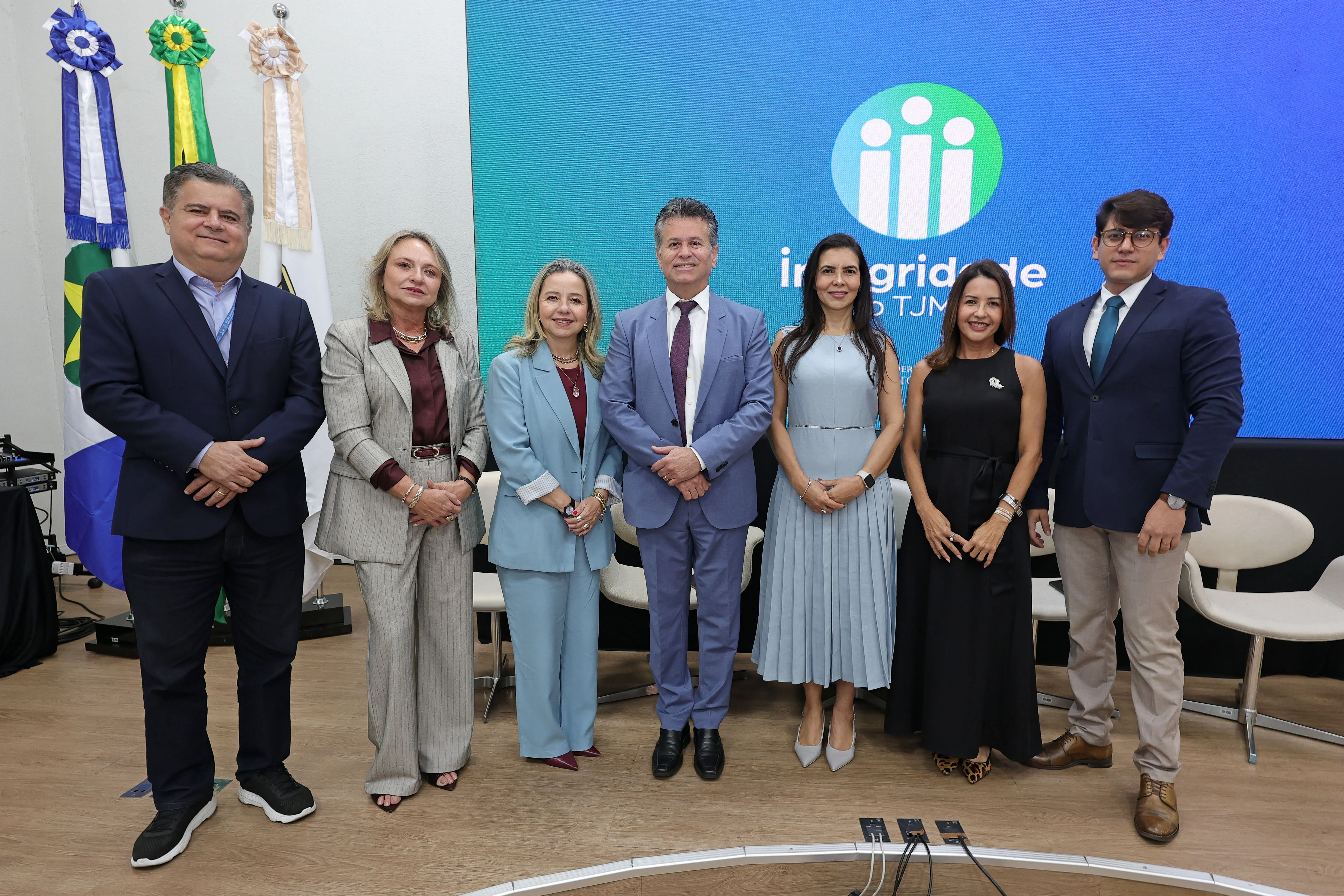 Sete autoridades sorriem alinhadas lado a lado no palco de um auditório, em frente a bandeiras oficiais e a um telão azul com a marca "Integridade do TJMT".