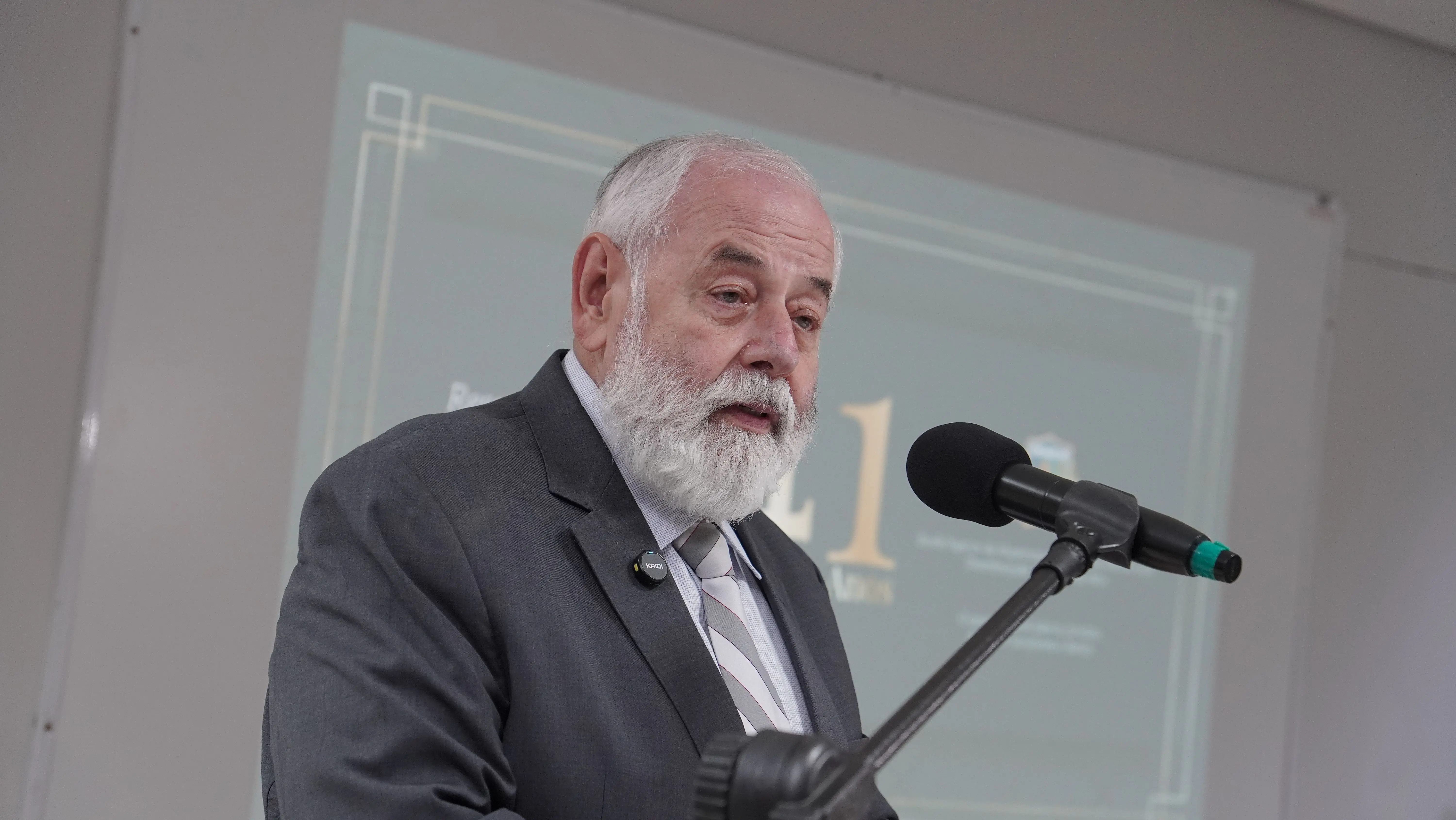 Homem idoso de barba branca discursa próximo a um microfone de pedestal. Ele veste terno cinza e gravata listrada. Ao fundo, uma tela de projeção exibe um slide esverdeado com o número 