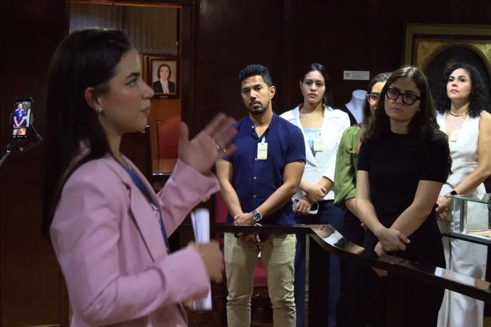 Foto horizontal em plano médio que mostra um grupo de estudantes de Direito observando uma mulher de blazer cor-de-rosa, que está à frente deles, falando. Eles estão no Espaço Memória do Tribunal de Justiça de Mato Grosso, local com paredes de madeira de jacarandá e iluminação baixa.