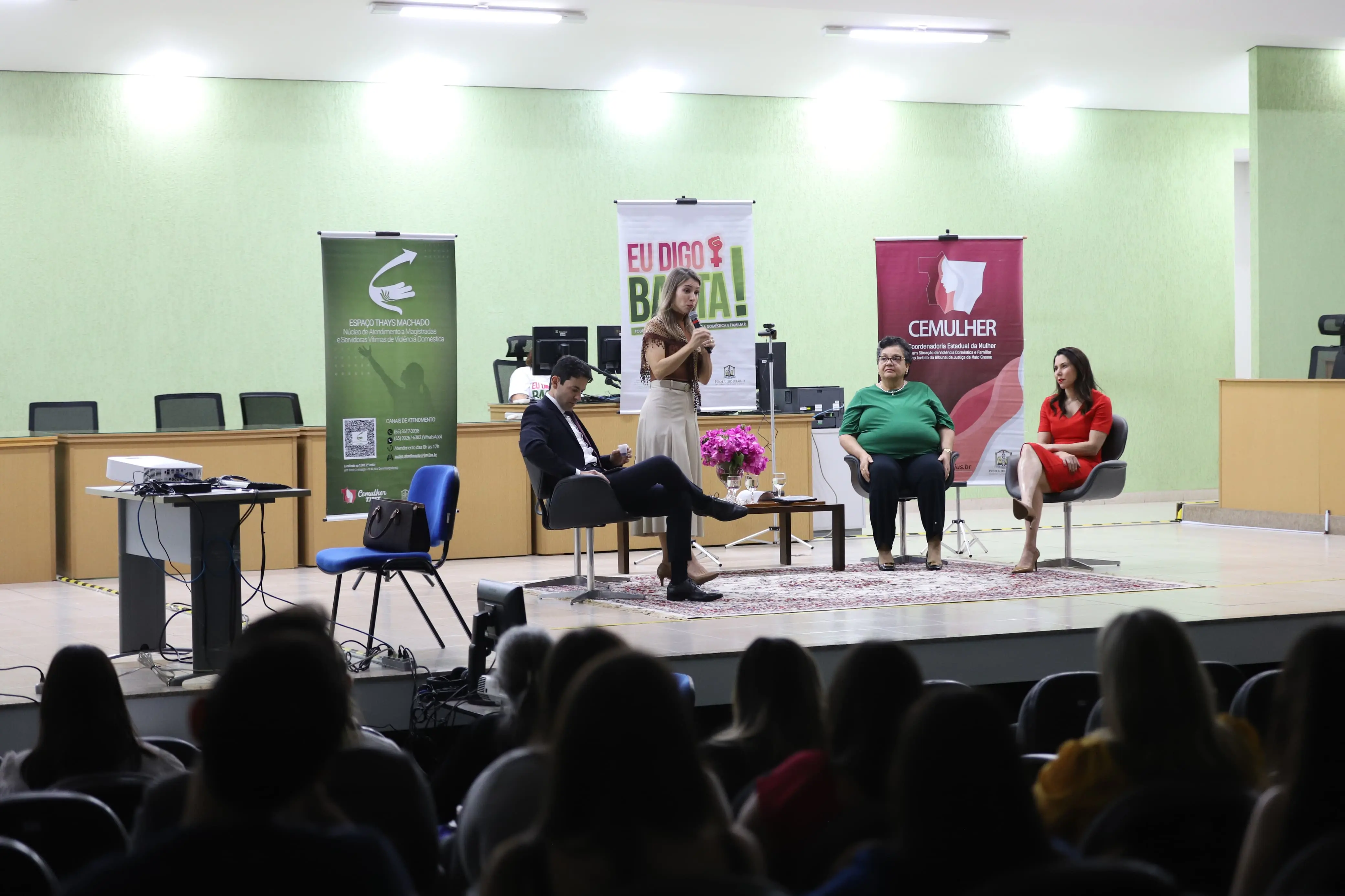 Evento em auditório reúne quatro pessoas no palco, sentadas em cadeiras modernas. Uma mulher fala ao microfone. Atrás, banners da campanha "Eu Digo Basta" e do CEMULHER. Público acompanha atento nas cadeiras.