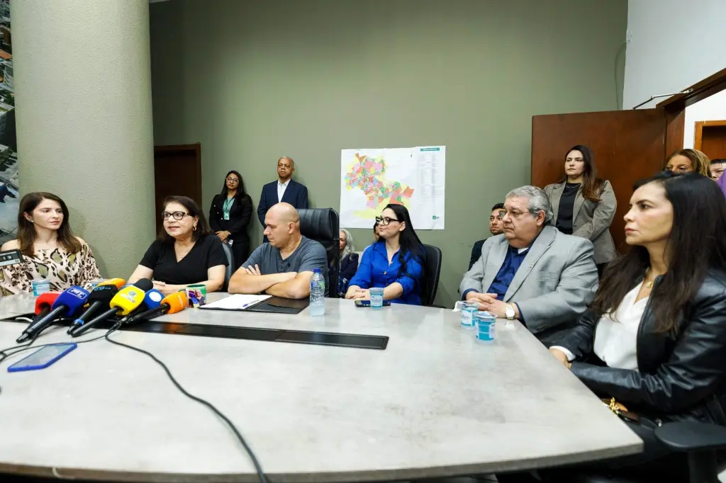 Autoridades estão sentadas ao redor de uma mesa oval em uma sala de reuniões. O prefeito está ao centro, ao lado das juízas, primeira-dama, procurador e promotora da Justiça