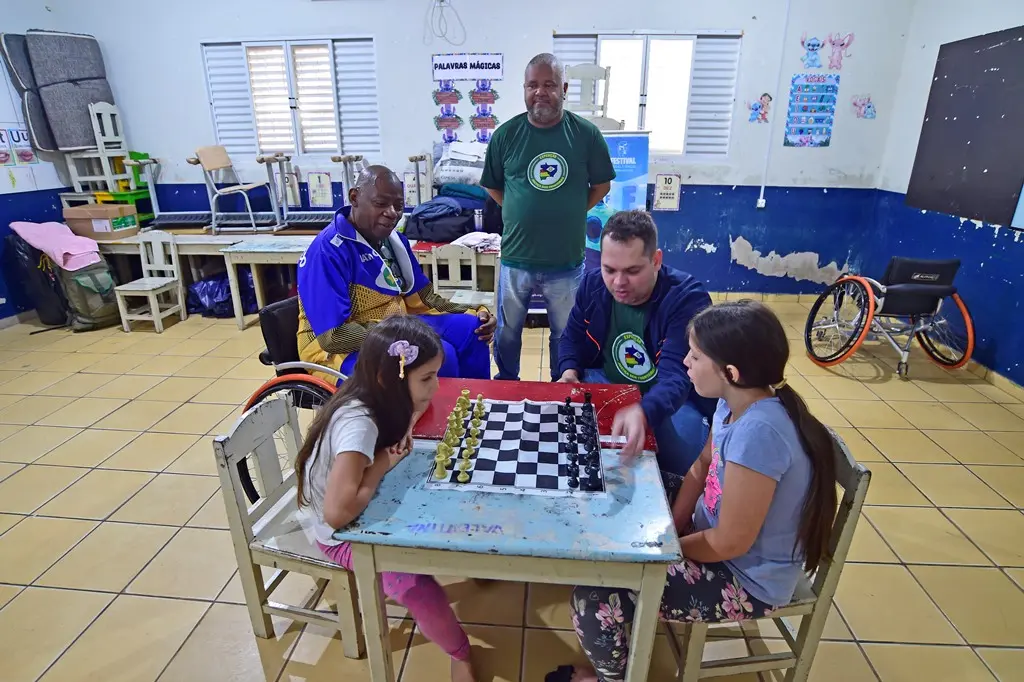 Duas meninas jogando xadrez em uma mesa no centro de uma sala de aula. Três servidores da Secretaria de Esporte assistem ao jogo. Um deles está sentado em uma cadeira de rodas.