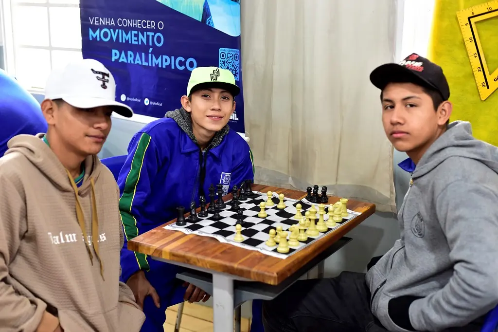 Três meninos usando moletom e boné posam para a foto, em volta de uma mesa com tabuleiro de xadrez.