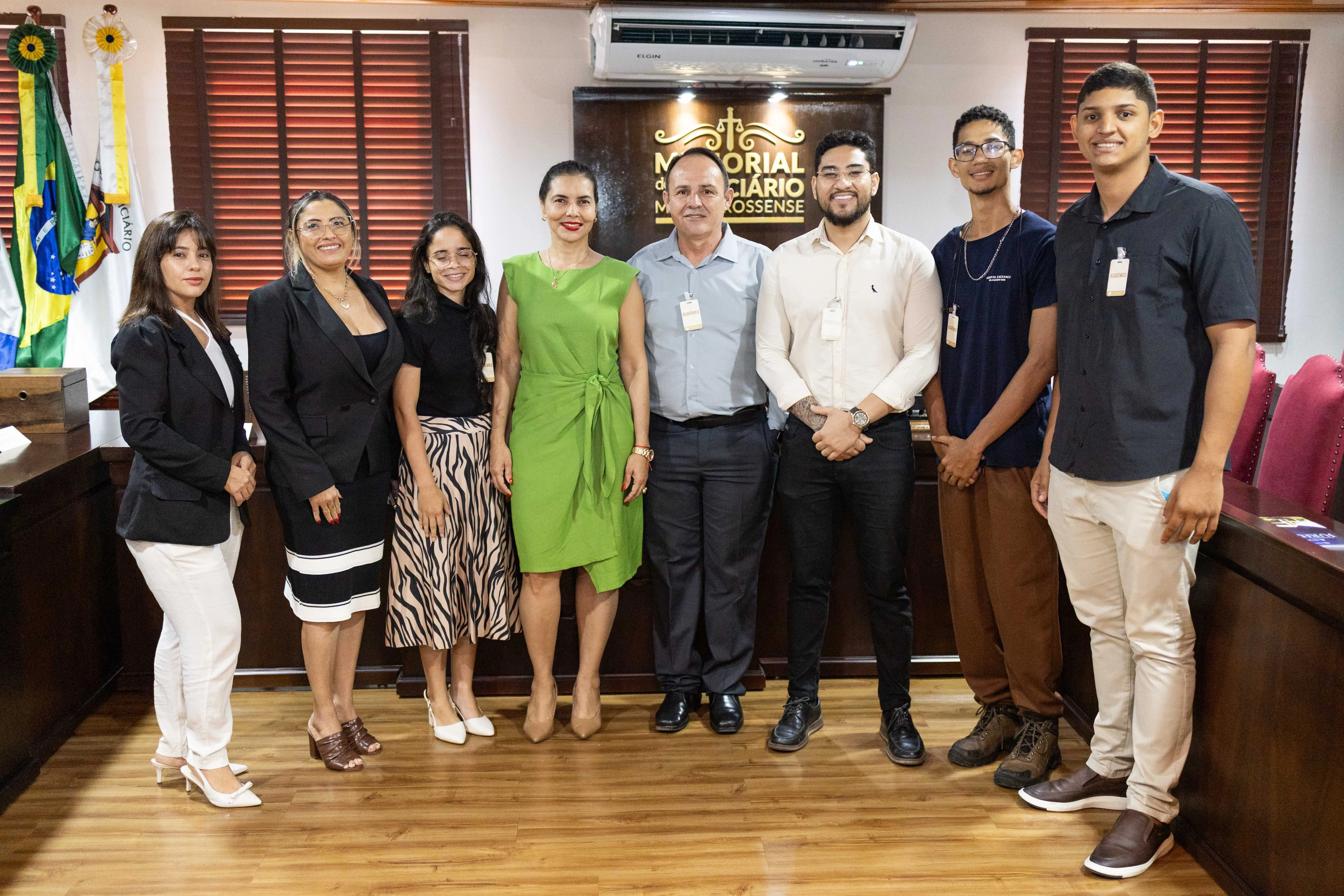 Oito pessoas posam sorrindo para foto em sala do Memorial do Judiciário Mato-grossense. Estão alinhadas lado a lado, com roupas formais. Ao fundo, há bandeiras, persianas e placa com o nome do local.