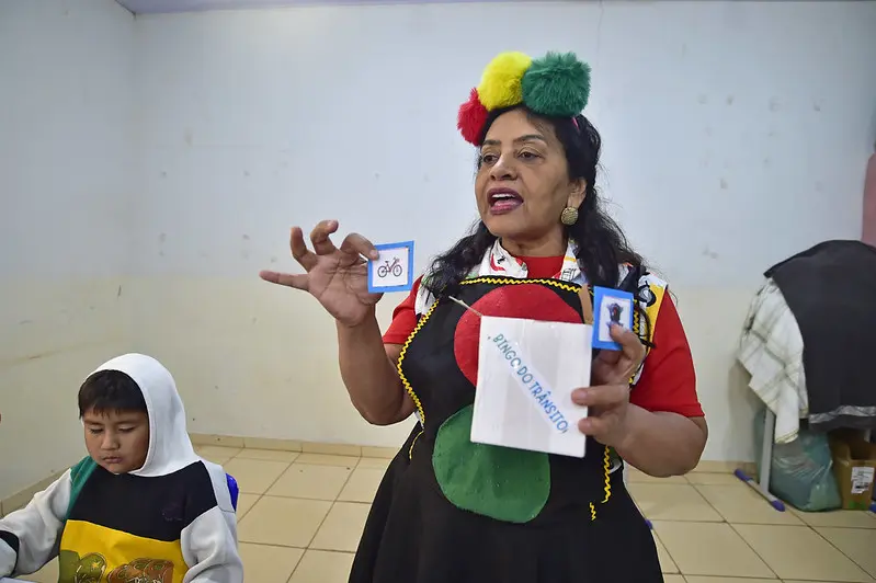Servidora do Detran profere palestra em uma sala de aula. Ela é uma mulher negra, de cabelos pretos, usando roupa colorida e pompons na cabeça. O figurino tem as cores vermelho, amarelo, verde e preto, simbolizando um semáforo. Há um menino sentado ao lado dela.