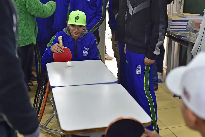 Meninos jogam tênis de mesa em cadeira de rodas em uma sala de aula. O menino segurando a raquete sorri. Há vários estudantes em volta.