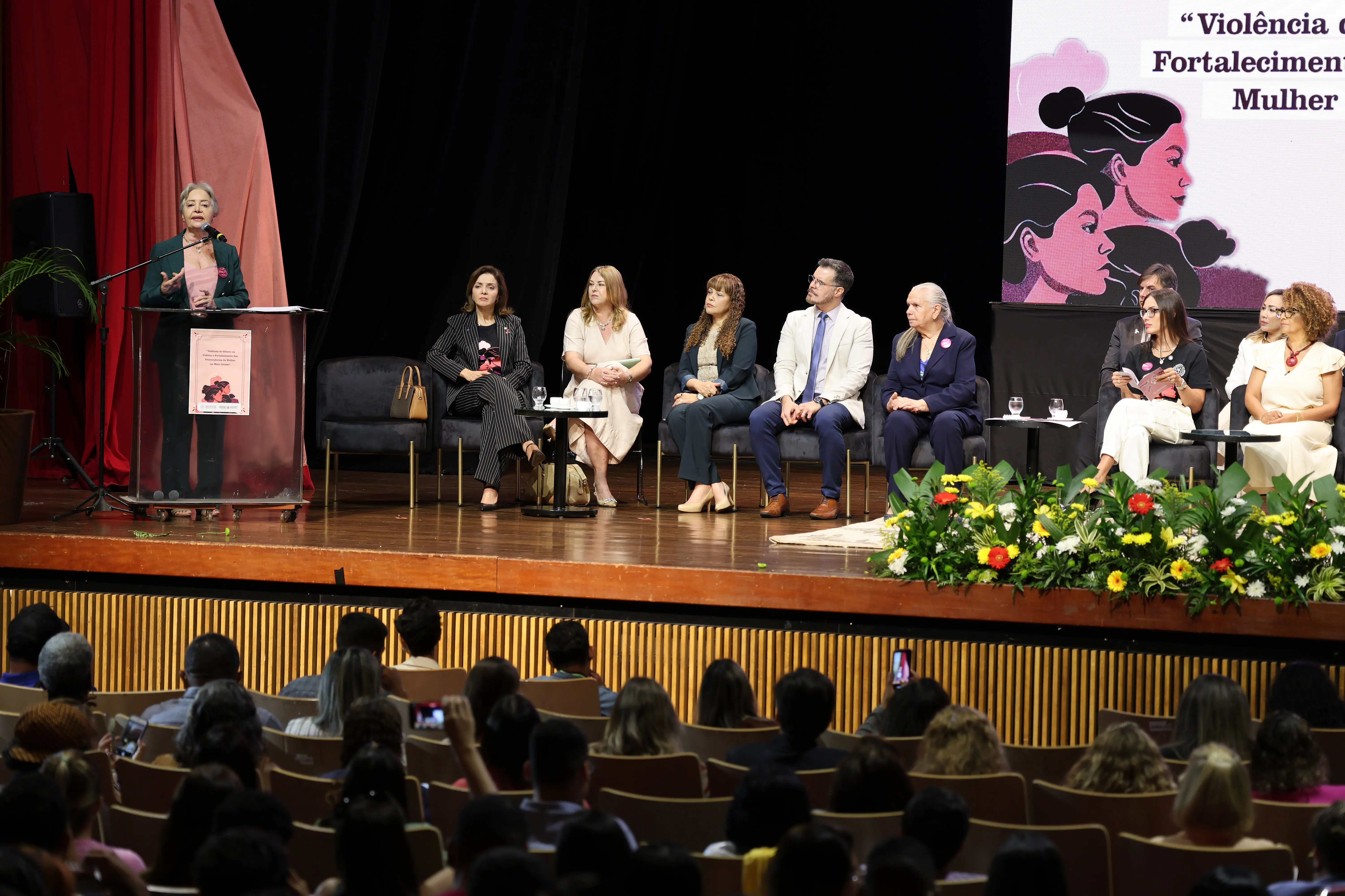 Auditório cheio acompanha painel do 1º Encontro da Procuradoria Especial da Mulher da ALMT. No palco, autoridades sentadas ou discursando debatem violência de gênero e fortalecimento da atuação feminina.