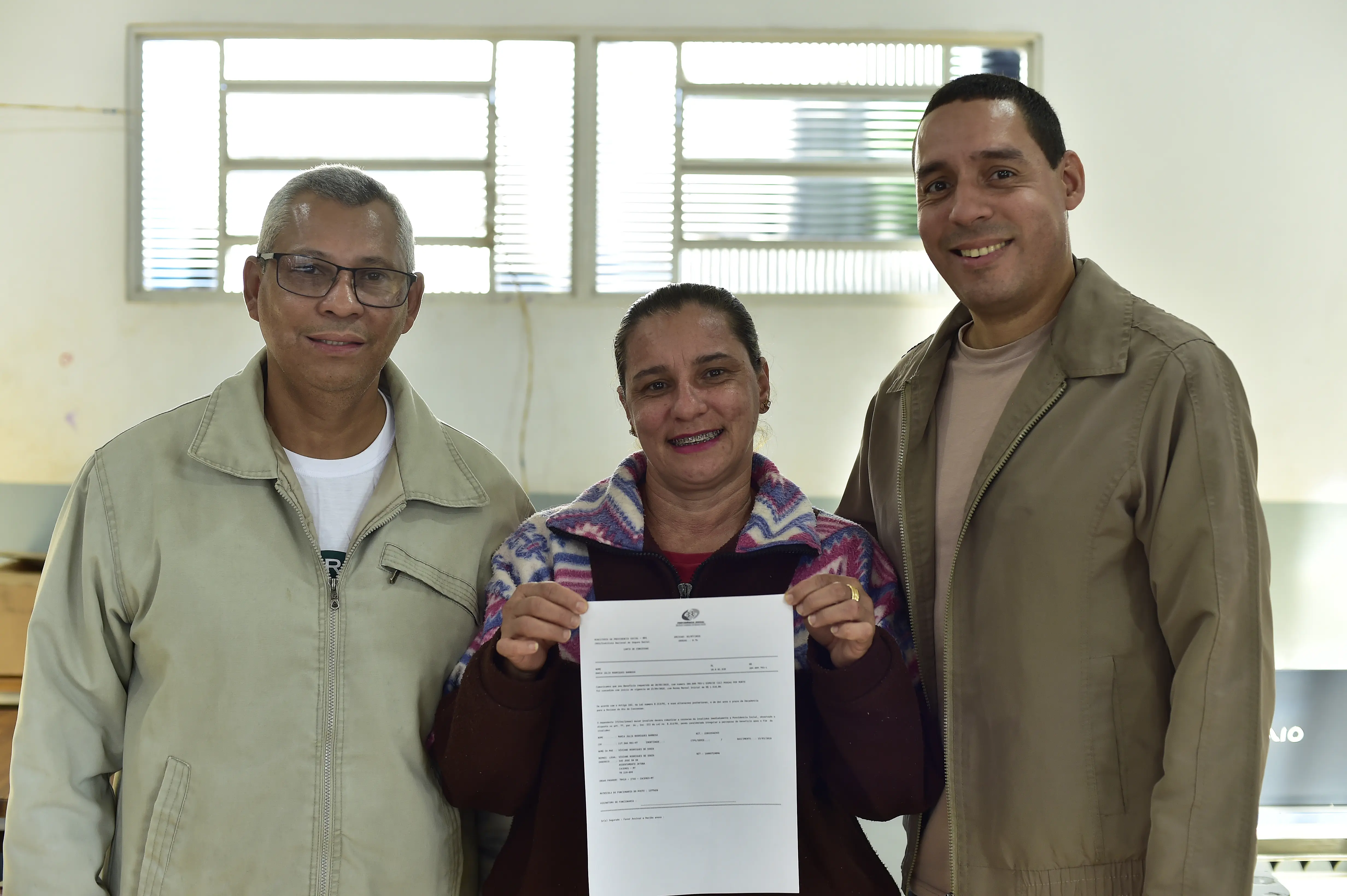 mulher aparece sorrindo ao centro, segurando o mesmo documento. Ela está acompanhada por dois homens, um de cada lado, também sorrindo. Ao fundo, vê-se uma janela e parte da sala com iluminação natural.