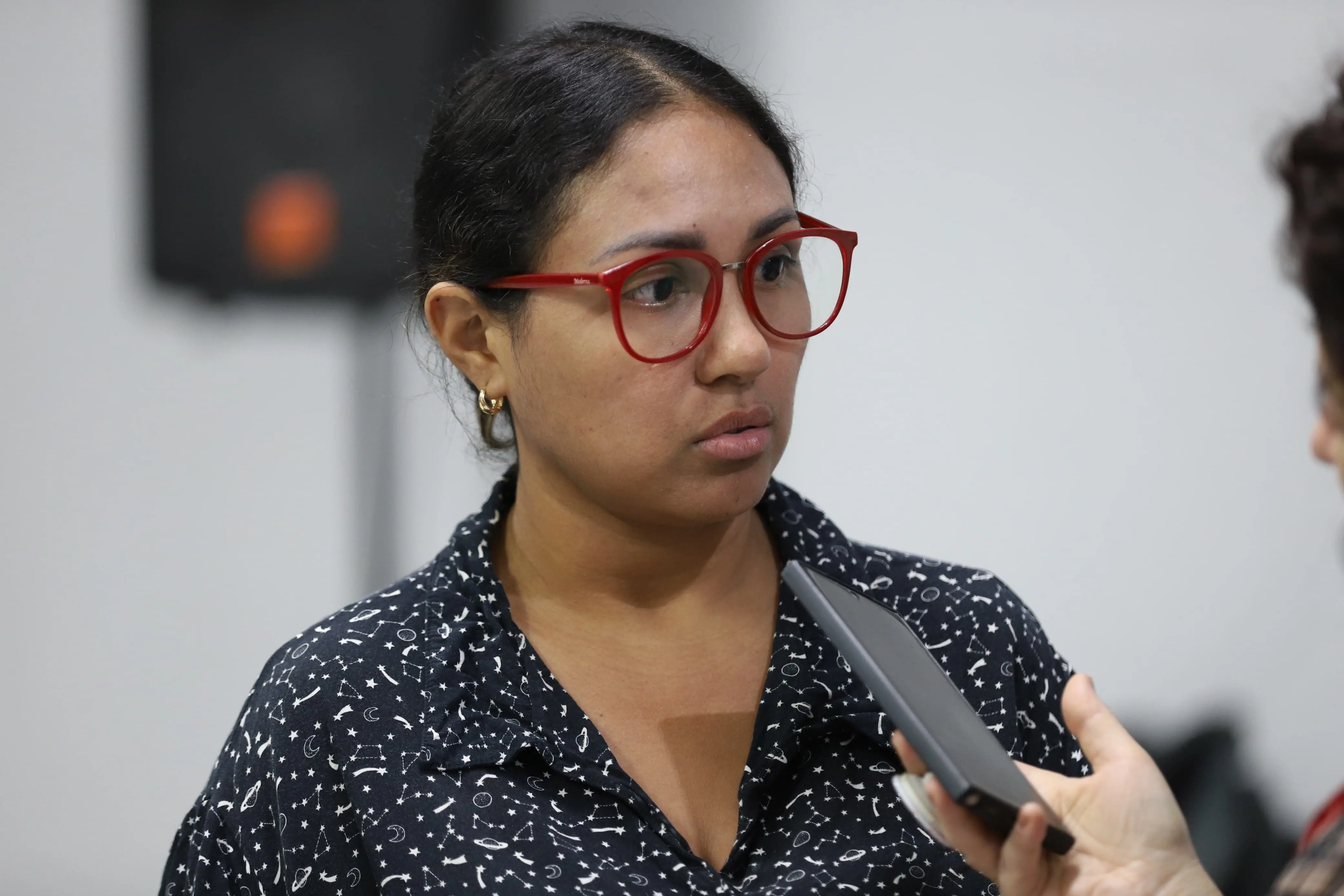 Mulher de óculos vermelhos e camisa escura estampada com astros. Ela está atenta, prestando depoimento. Detalhes visuais que tornam a informação acessível a todos.