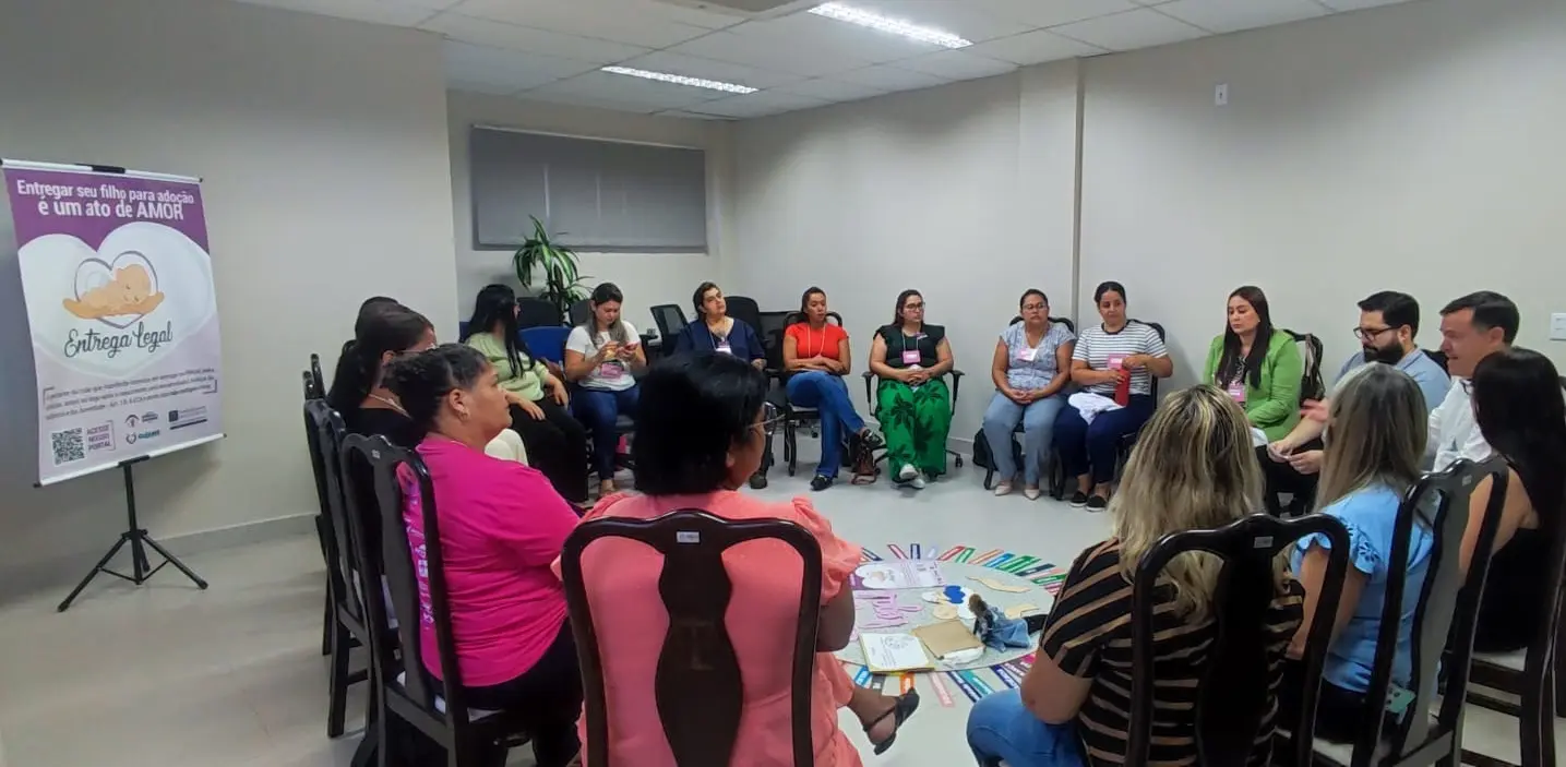 Um grupo de cerca de 15 pessoas sentadas em círculo em uma sala. Um banner roxo com o texto "Entregar seu filho para adoção é um ato de AMOR" está à esquerda.