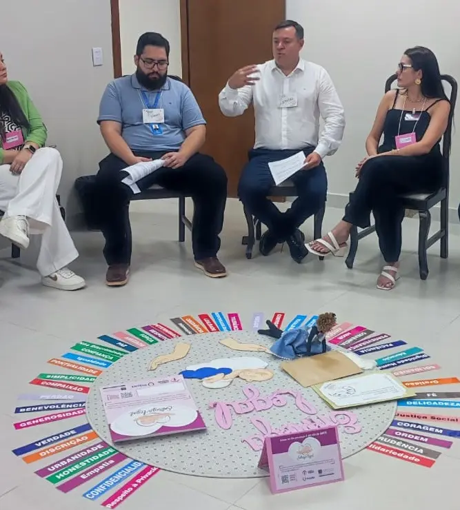 Grupo de pessoas sentadas em círculo no chão em torno de um diagrama com palavras como "Paz", "Coragem" e "Empatia". Um homem de camisa branca gesticula enquanto fala.