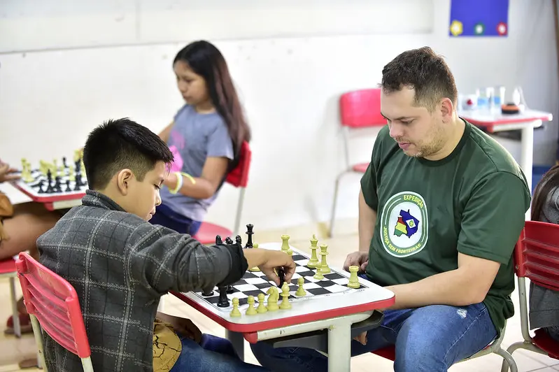 Um menino e um homem, usando camiseta verde da Justiça Sem Fronteiras, jogam xadrez, em uma sala de aula. Ao fundo, é possível ver outra dupla jogando xadrez em outra mesa.