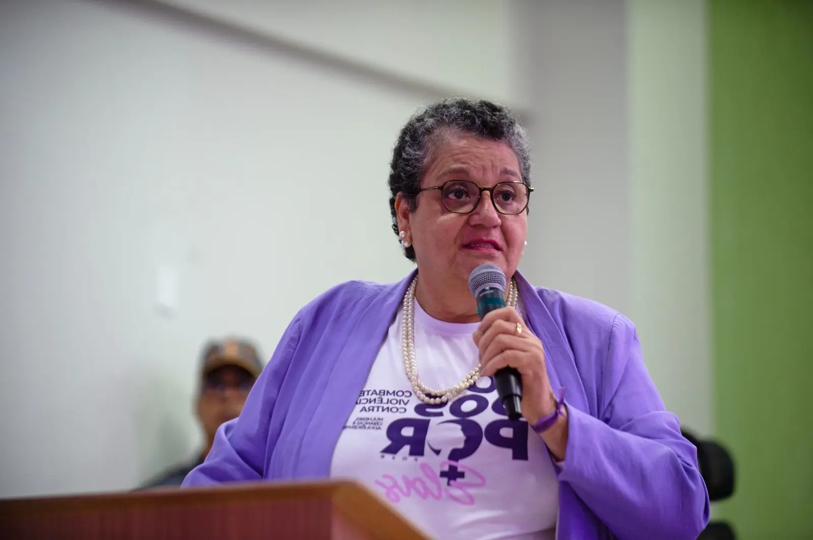 JuízaMaria Mazarelo fala ao microfone. Ela está atrás do púlpito. Usa camiseta branca com o nome do evento escrito em roxo. Usa um blaser lilás. Ela é uma mulher branca, de cabelos curtos com mechas grisalhas e usa óculos de grau e colar de pérolas