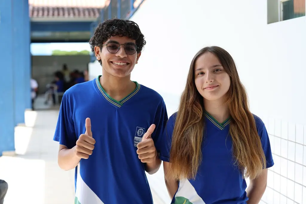 Foto horizontal em plano médio que mostra os estudantes Gabriel Henrique e Evelyn sorrindo para a foto, no pátio da escola. Gabriel faz sinal de joia com as duas mãos. Eles estão usando uniforme escolar azul. Gabriel é negro e usa óculos de grau solar e Evelyn é branca com longos cabelos loiros.