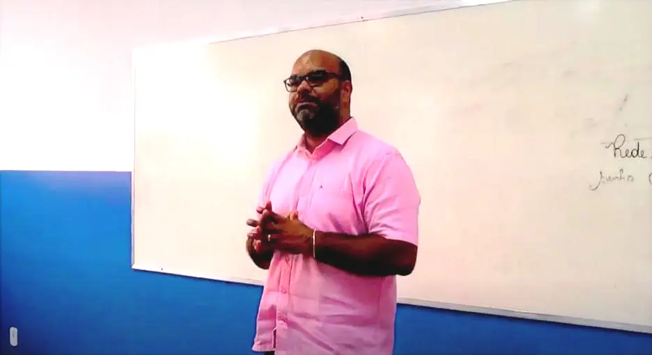 Homem de camisa rosa, óculos e barba, está de pé diante de um quadro branco