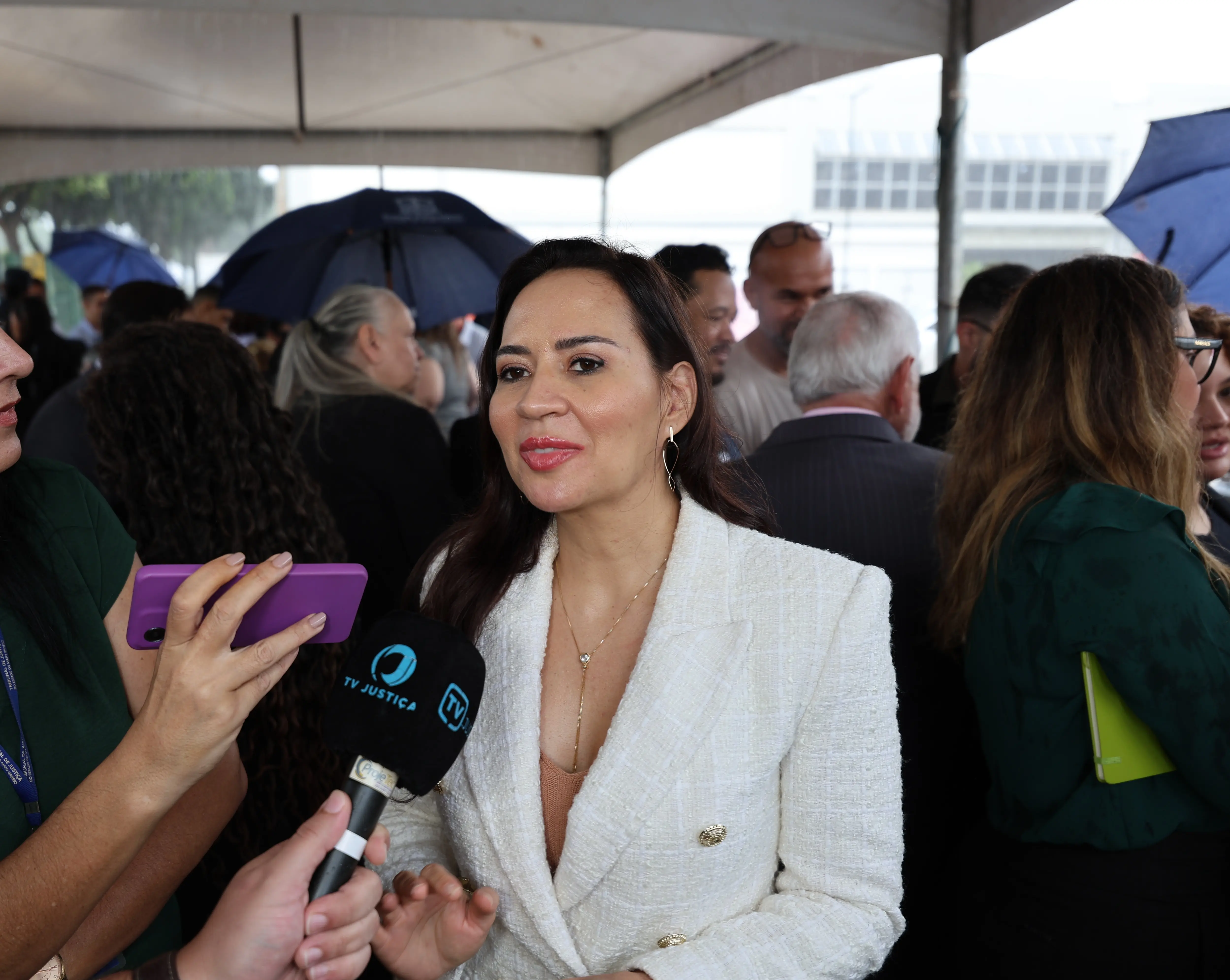 Juíza Tatyana Lopes, mulher de cabelos castanhos longos, veste blazer branco texturizado. Ela fala lateralmente enquanto segura um microfone da TV Justiça. Ambiente externo sob tenda com pessoas ao fundo.