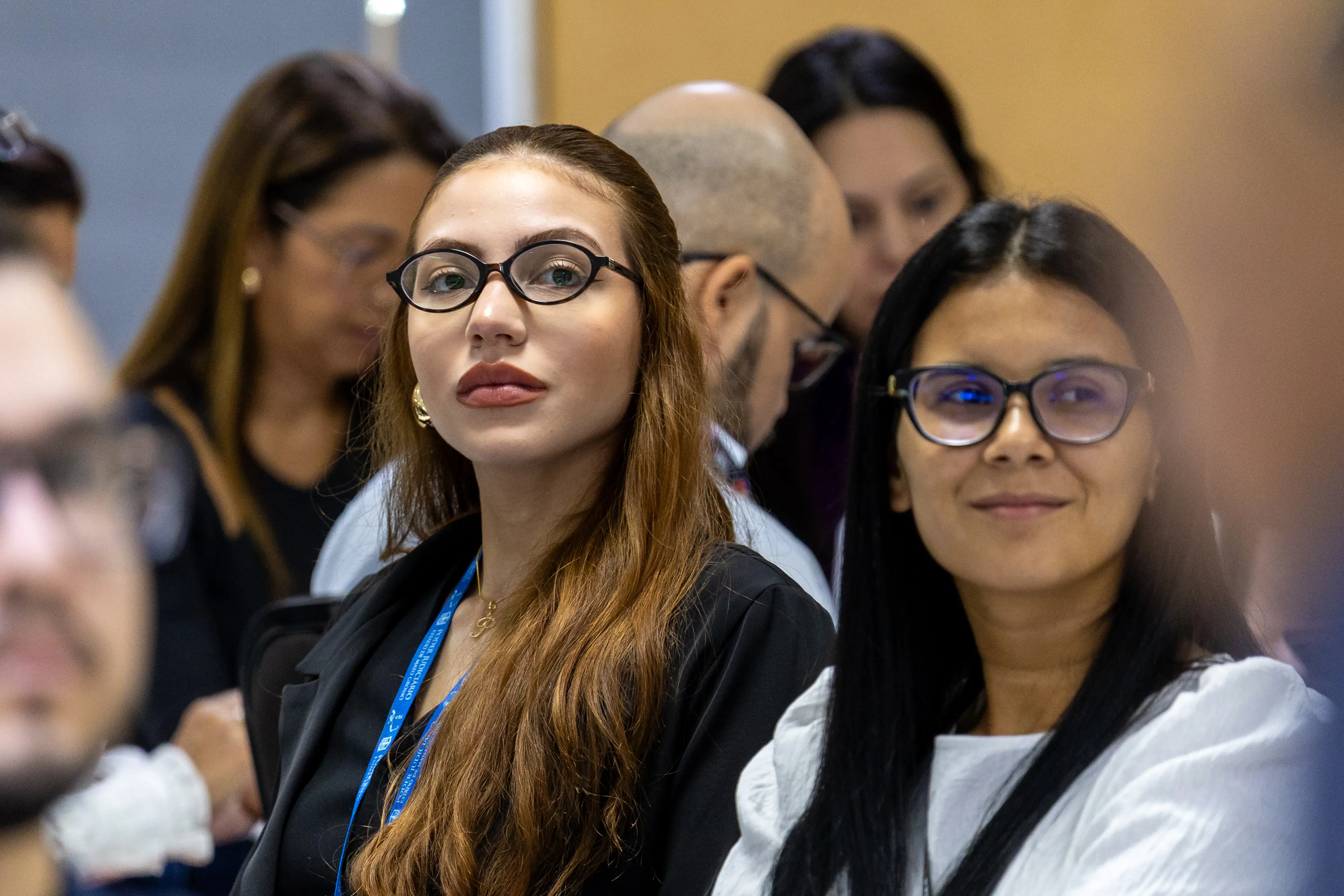 Duas mulheres em primeiro plano observam a apresentação. Ambas usam óculos e crachá institucional. Ao fundo, outros participantes mantêm foco nas discussões do encontro.