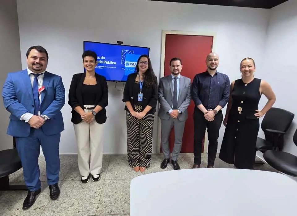 Seis pessoas sorriem em uma sala de reunião. Ao fundo, uma TV exibe o logo 