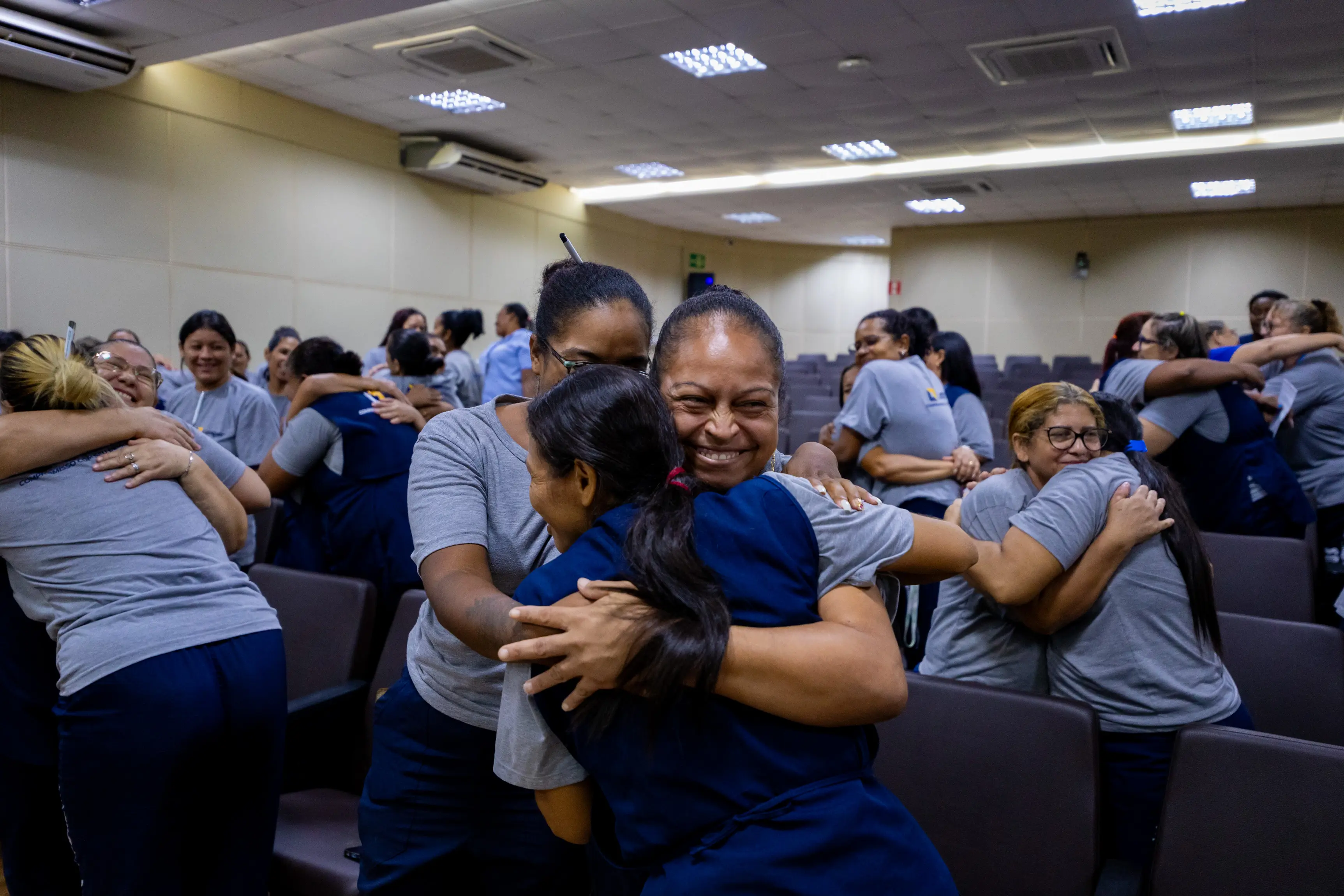 Participantes do evento aparecem abraçados em grupo dentro do auditório. Pessoas vestidas de azul demonstram acolhimento e emoção durante atividade de integração realizada com os participantes.