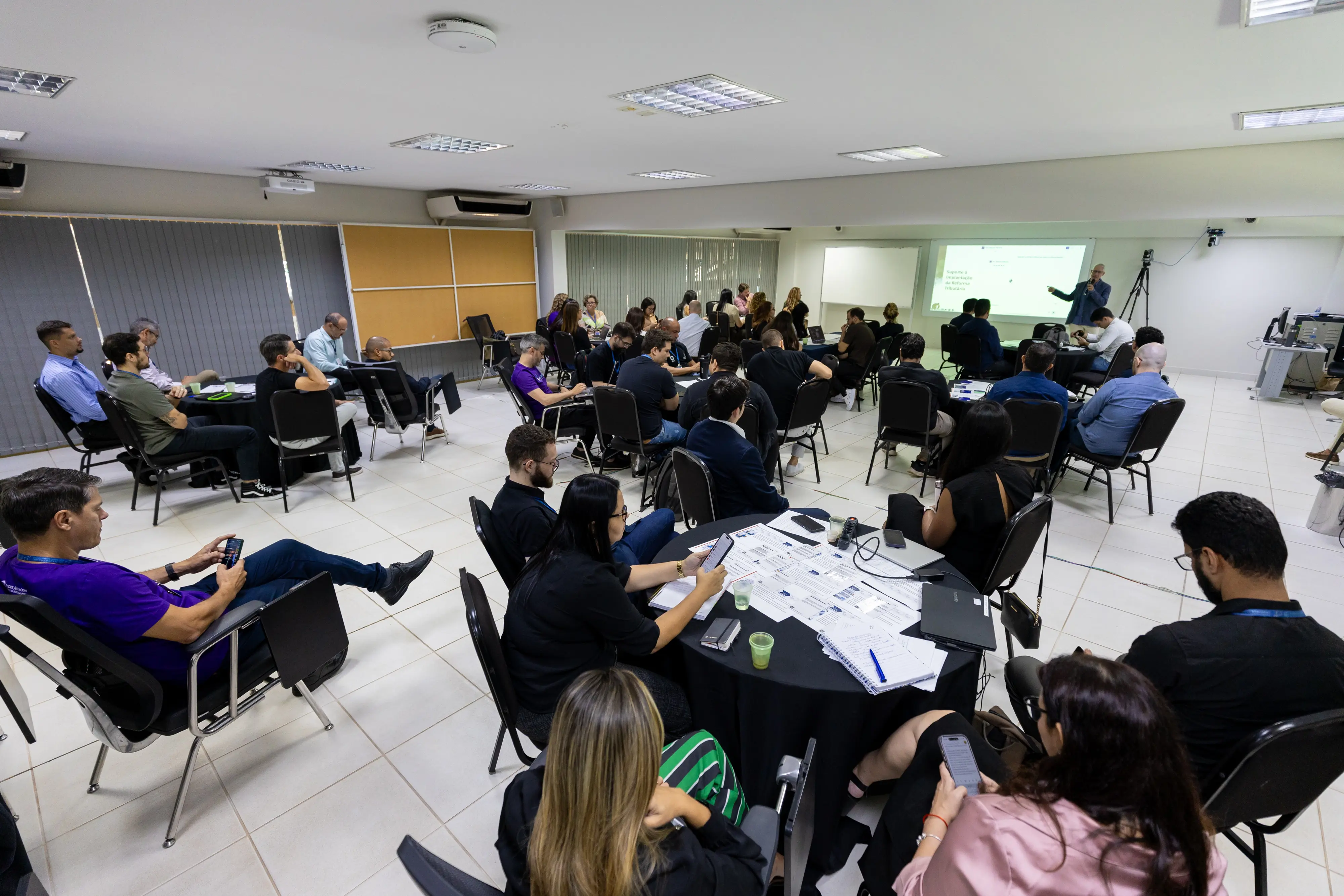 Sala ampla com participantes distribuídos em mesas redondas, atentos à apresentação projetada. Um palestrante aponta para o slide enquanto o público utiliza notebooks e celulares em ambiente corporativo iluminado.