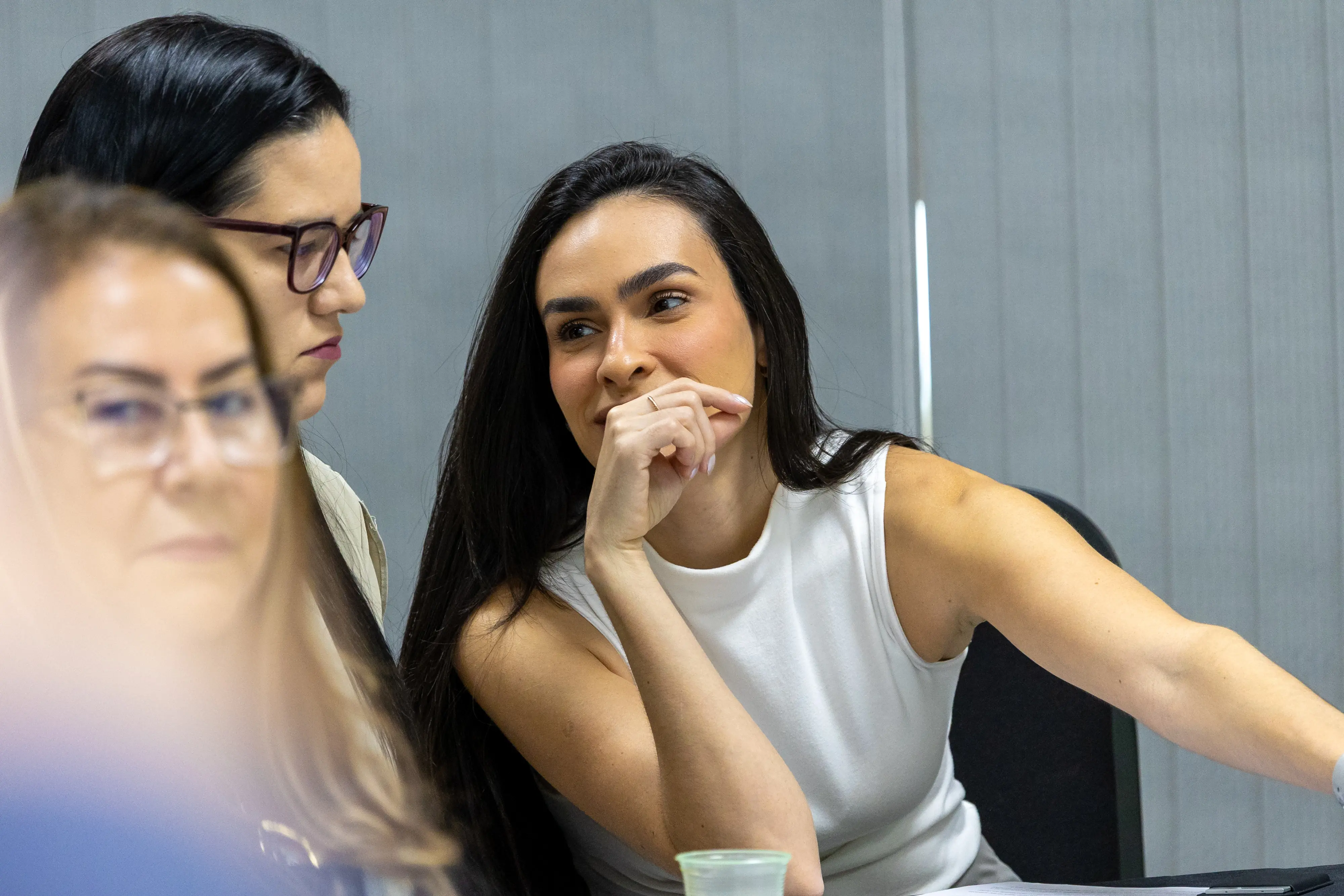 Grupo de mulheres interage durante atividade. Uma delas apoia a mão no rosto enquanto conversa com colega. Expressões demonstram concentração e troca de ideias.
