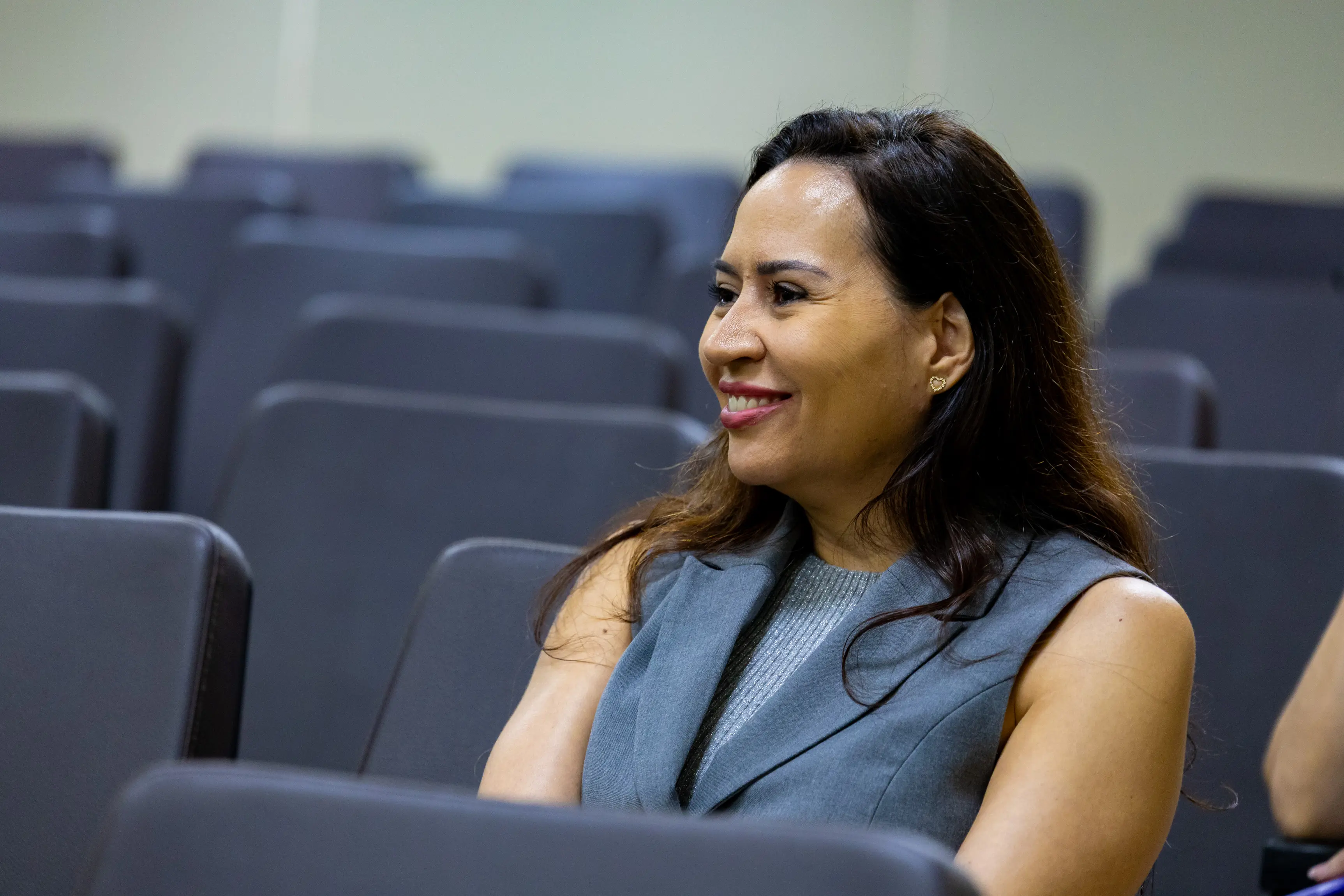 Mulher de cabelos longos e blusa cinza está sentada em cadeira do auditório e sorri enquanto observa a atividade. O ambiente possui fileiras de assentos estofados e iluminação clara no espaço.