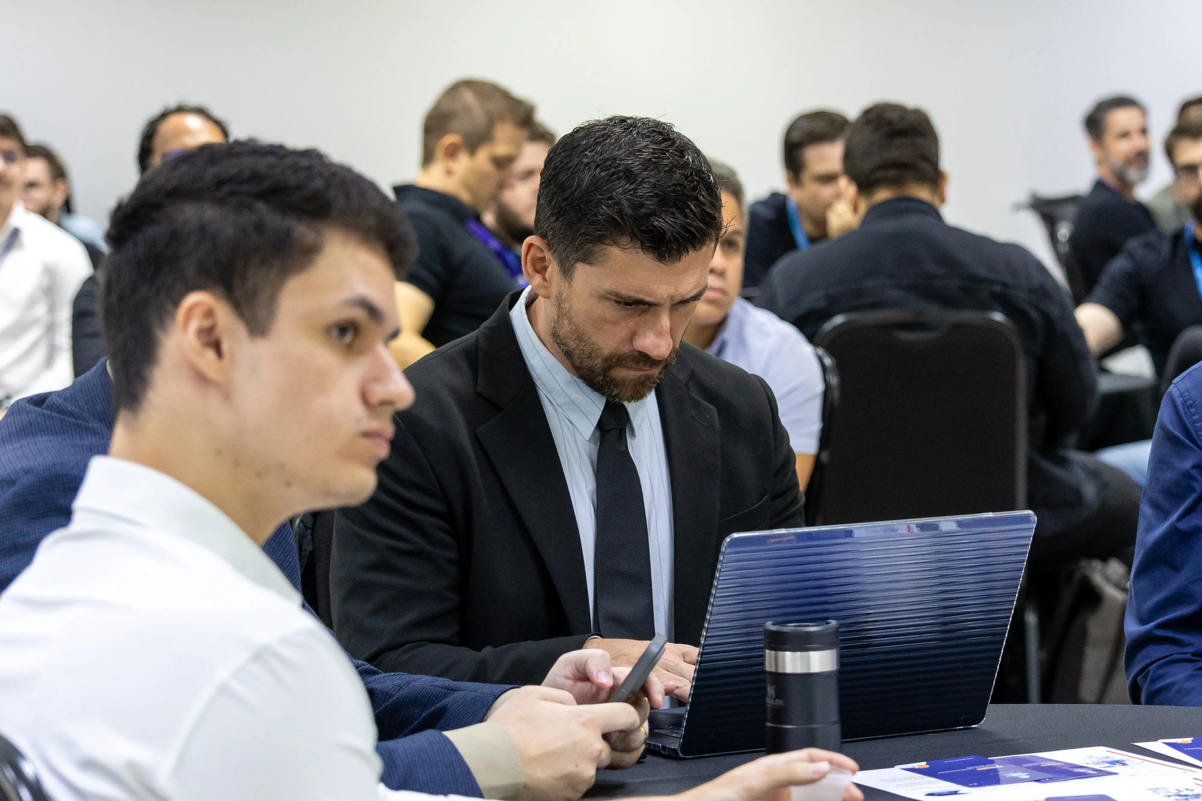 Participantes sentados acompanham atividade prática. Homem de terno digita no notebook enquanto colega ao lado consulta celular. Ao fundo, grupo observa atentamente a dinâmica proposta.