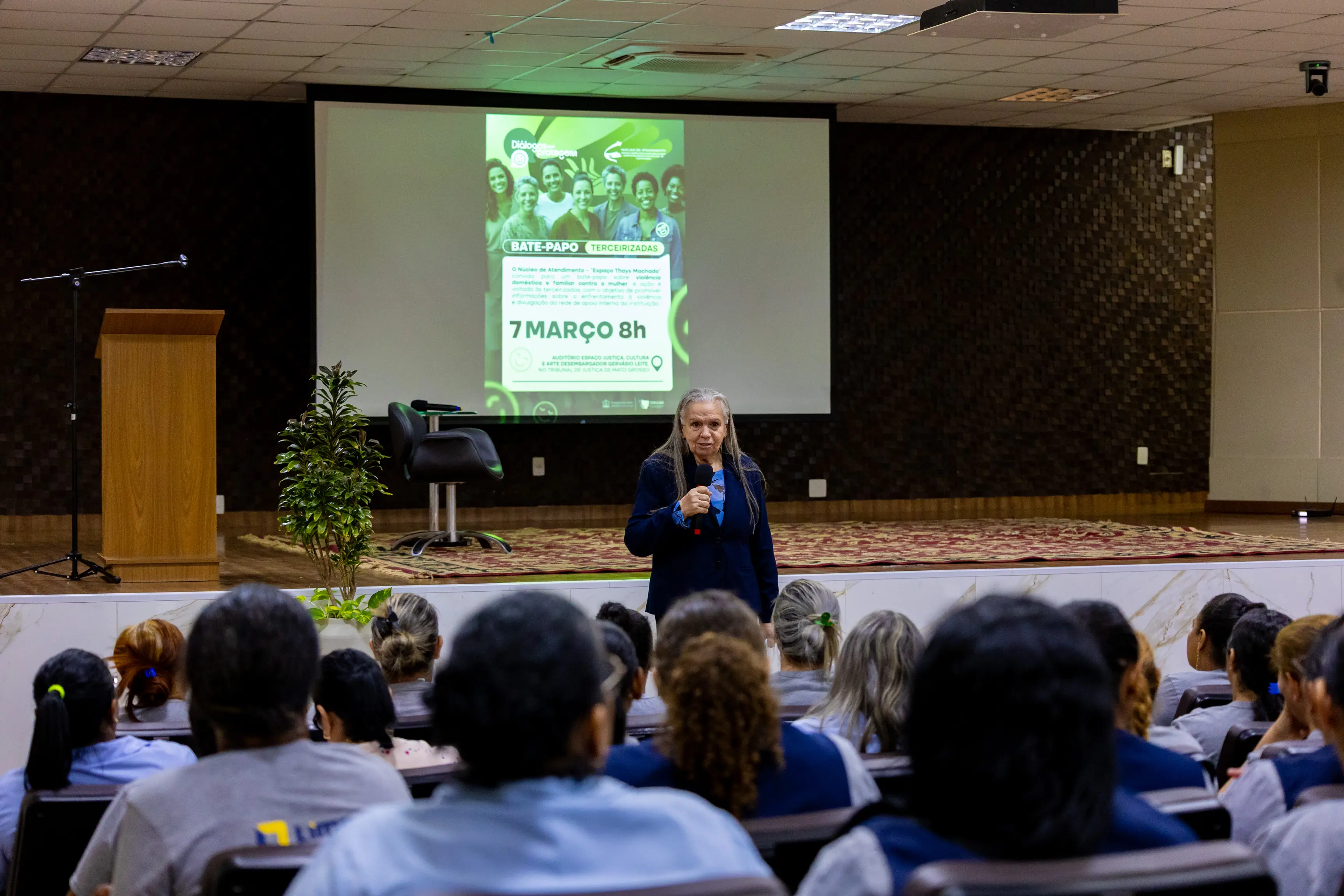Auditório visto de frente mostra desembargadora Maria Erotides alando ao microfone diante de plateia numerosa. O telão atrás exibe slide do evento, reforçando o ambiente de palestra institucional.