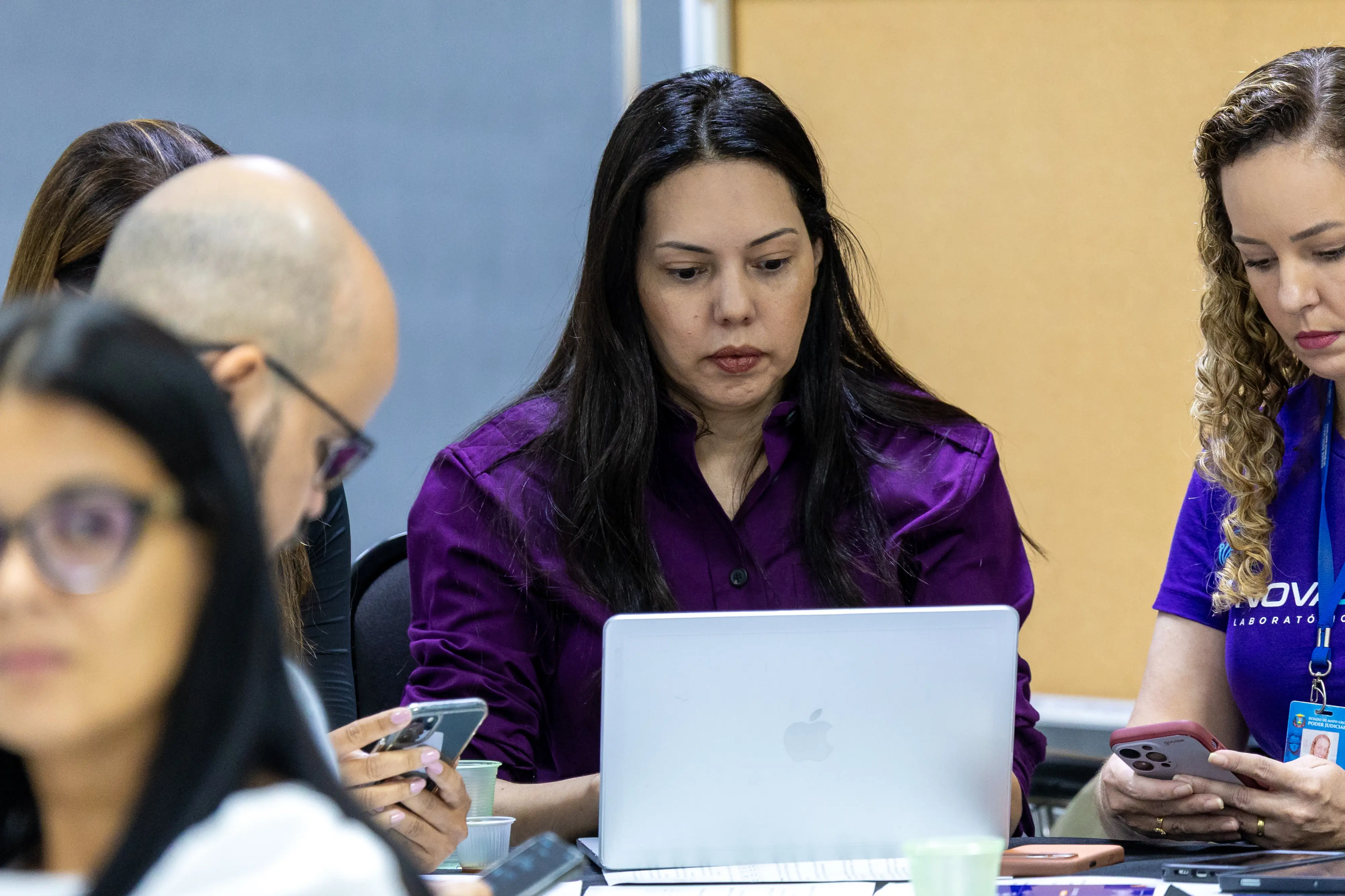 Participantes analisam conteúdos em mesa compartilhada. Mulher ao centro utiliza notebook enquanto outras consultam celulares. O clima é de estudo e colaboração.