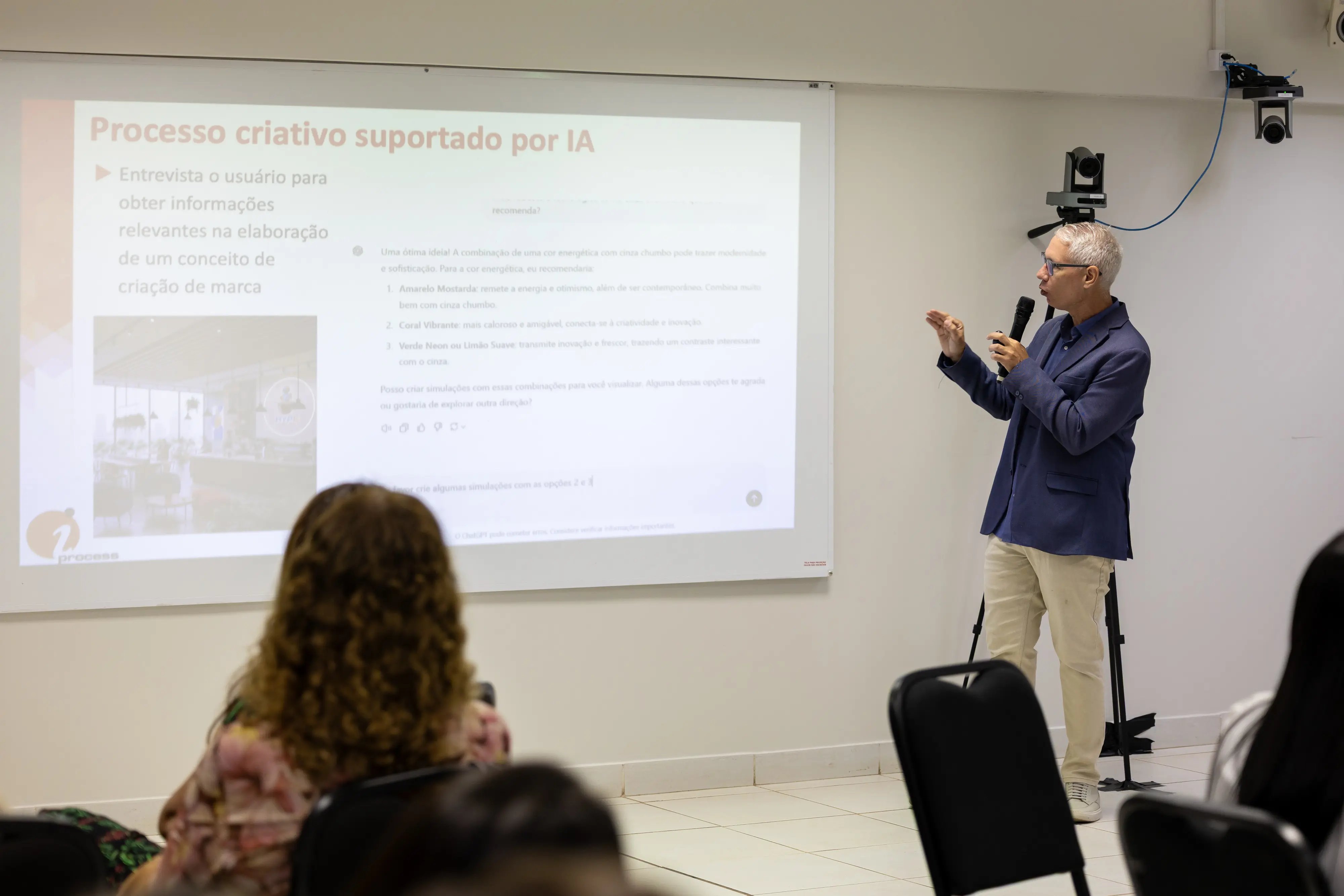 Homem de blazer azul apresenta conteúdo sobre processo criativo com apoio de IA. Ele segura microfone e gesticula diante de telão com texto e imagens, em sala de treinamento estruturada.