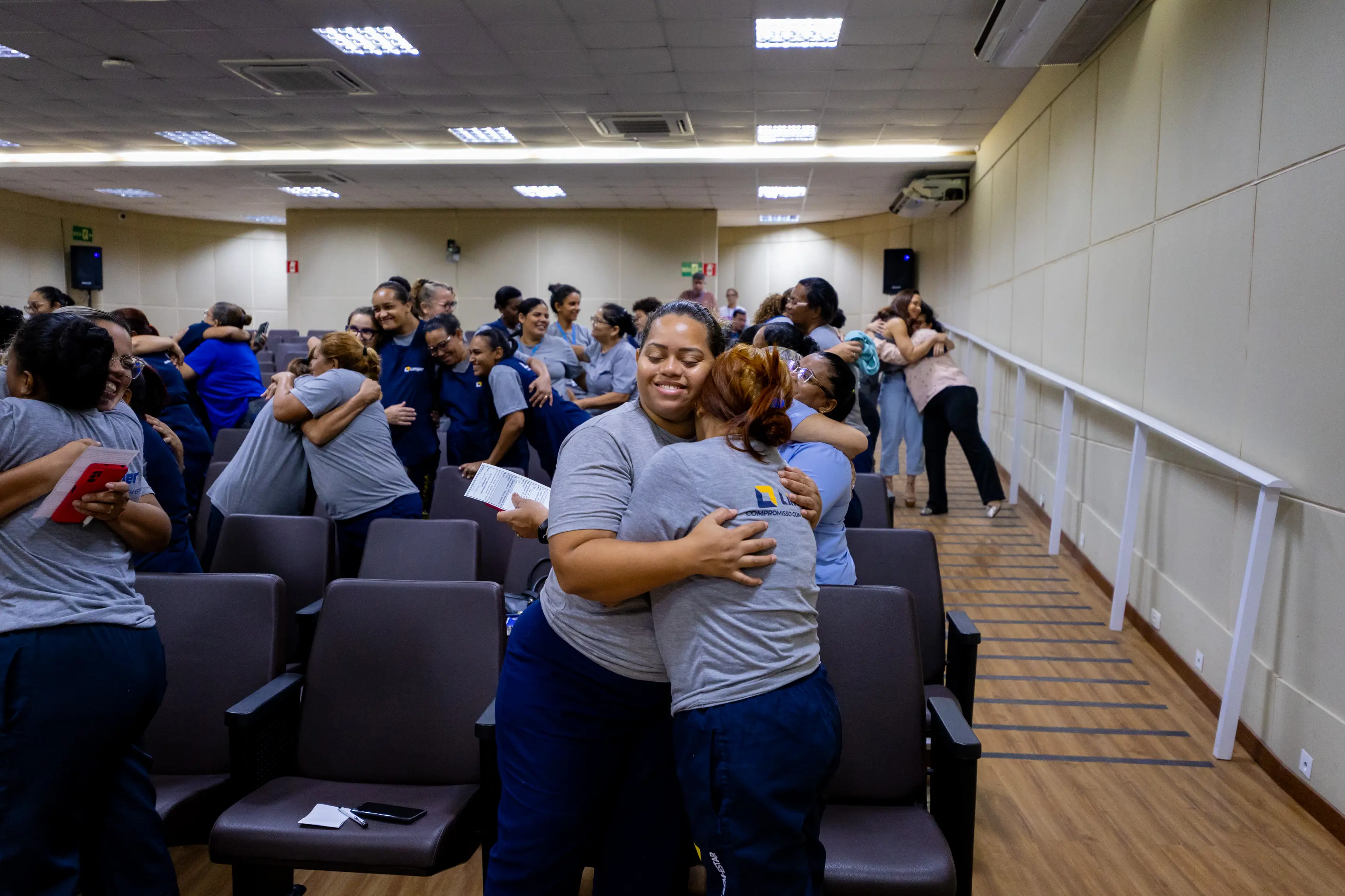 No auditório, duas participantes se abraçam no centro da imagem enquanto outras pessoas também realizam a mesma dinâmica nas fileiras ao redor. O momento demonstra interação, empatia e integração entre o grupo.