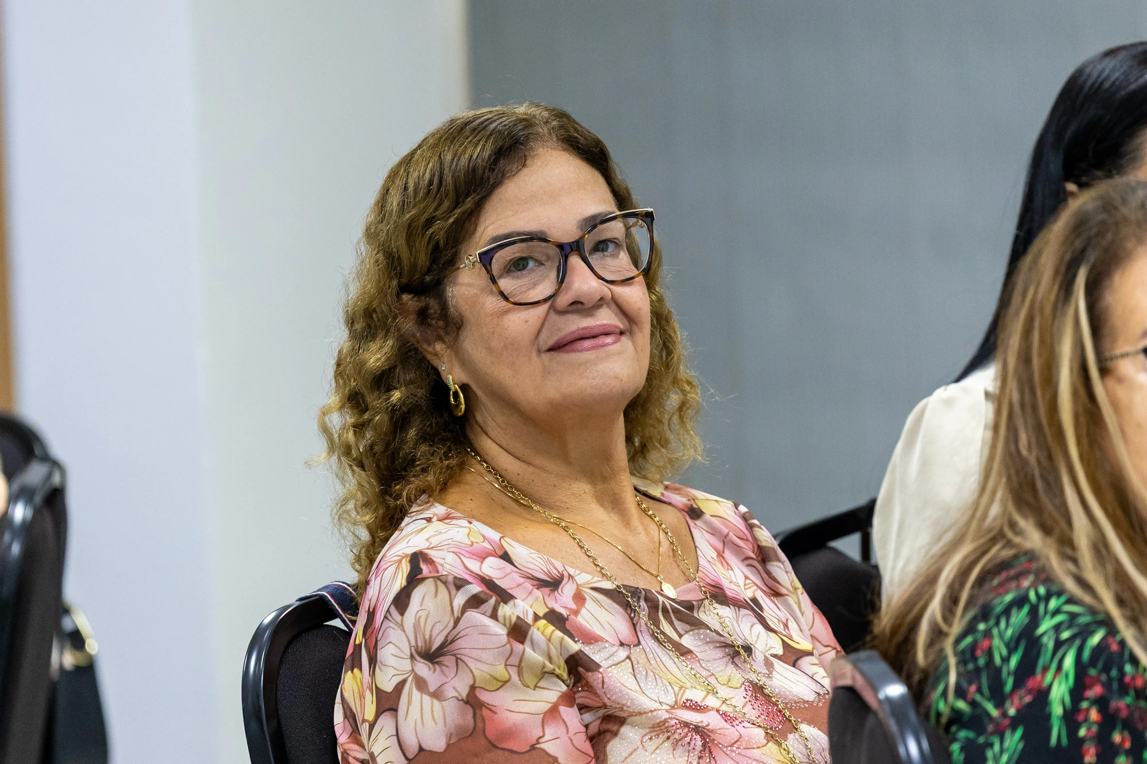 Mulher de óculos, com blusa floral, está sentada e olha em direção à câmera. O ambiente mostra colegas ao redor, compondo cenário de capacitação profissional.