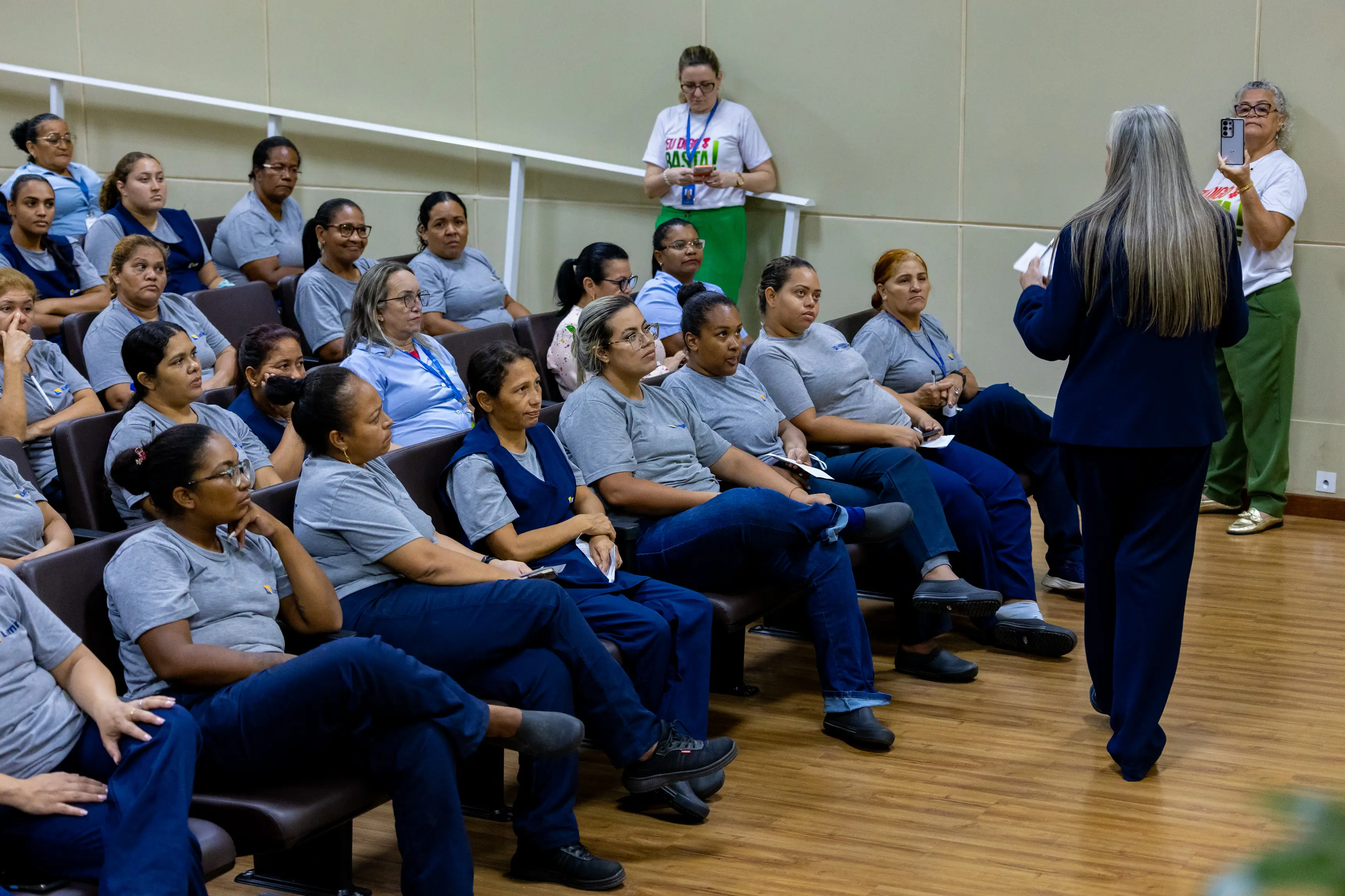 Participantes do evento estão sentados em cadeiras do auditório, usando camisetas azuis e acompanhando atentamente uma facilitadora que fala em pé à frente do grupo. O momento mostra interação e atenção coletiva.