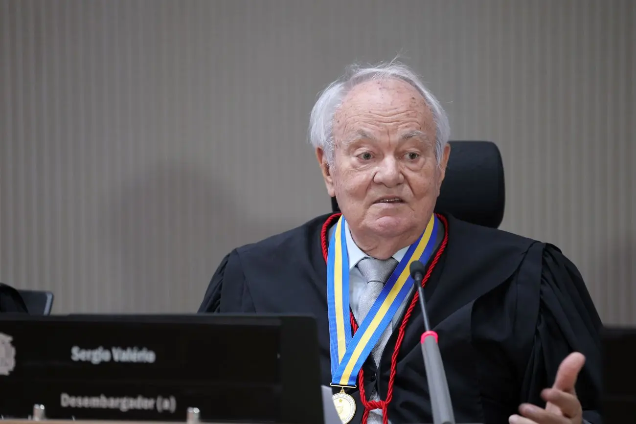 Foto horizontal que mostra o desembargador Sérgio Valerio sentado e falando ao microfone, gesticulando. Ele usa toga e medalha do mérito judiciário.