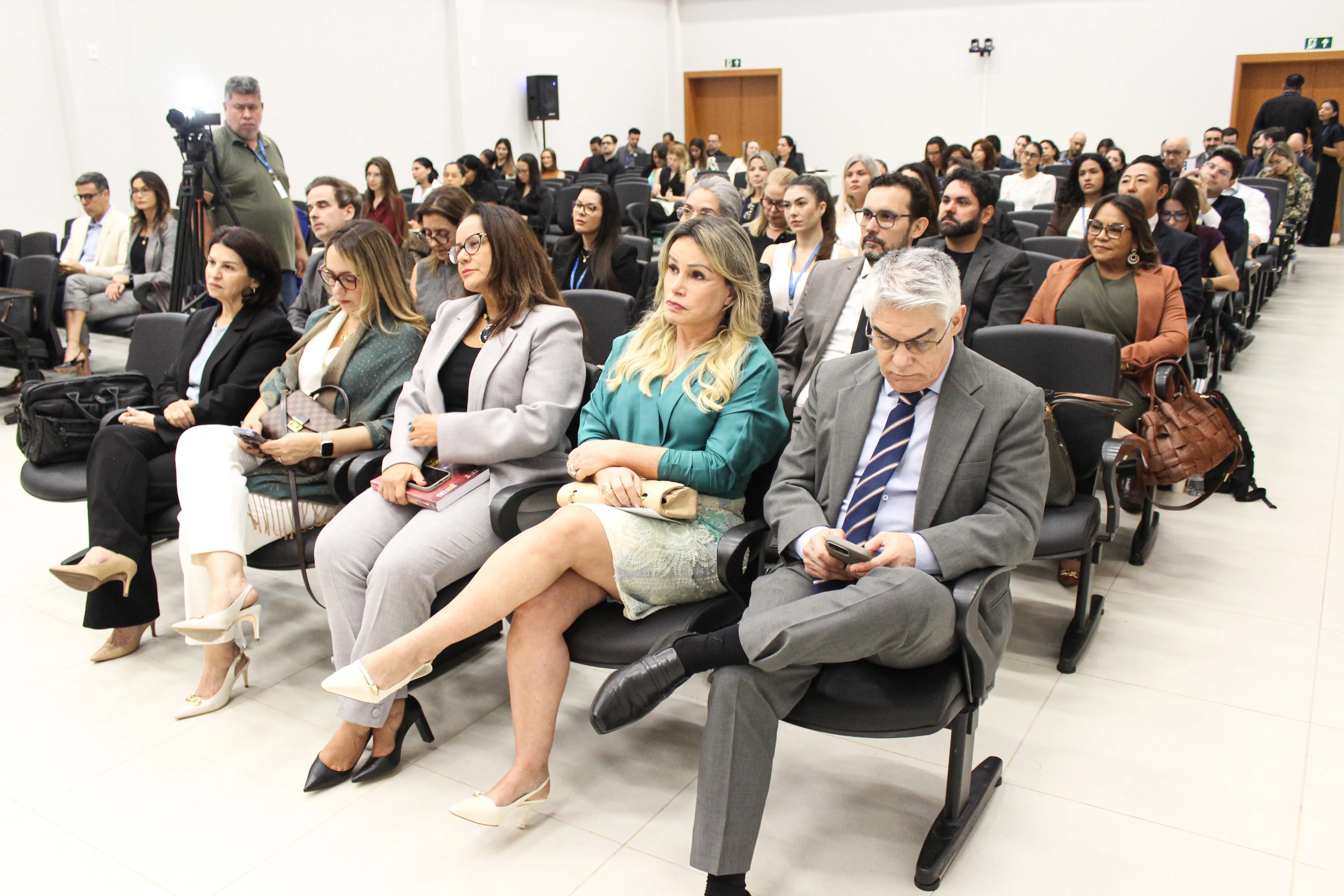 Plateia sentada em auditório durante evento formal. Em primeiro plano, homens e mulheres vestindo trajes formais e sociais acompanham a ocasião dispostos em fileiras de poltronas pretas.